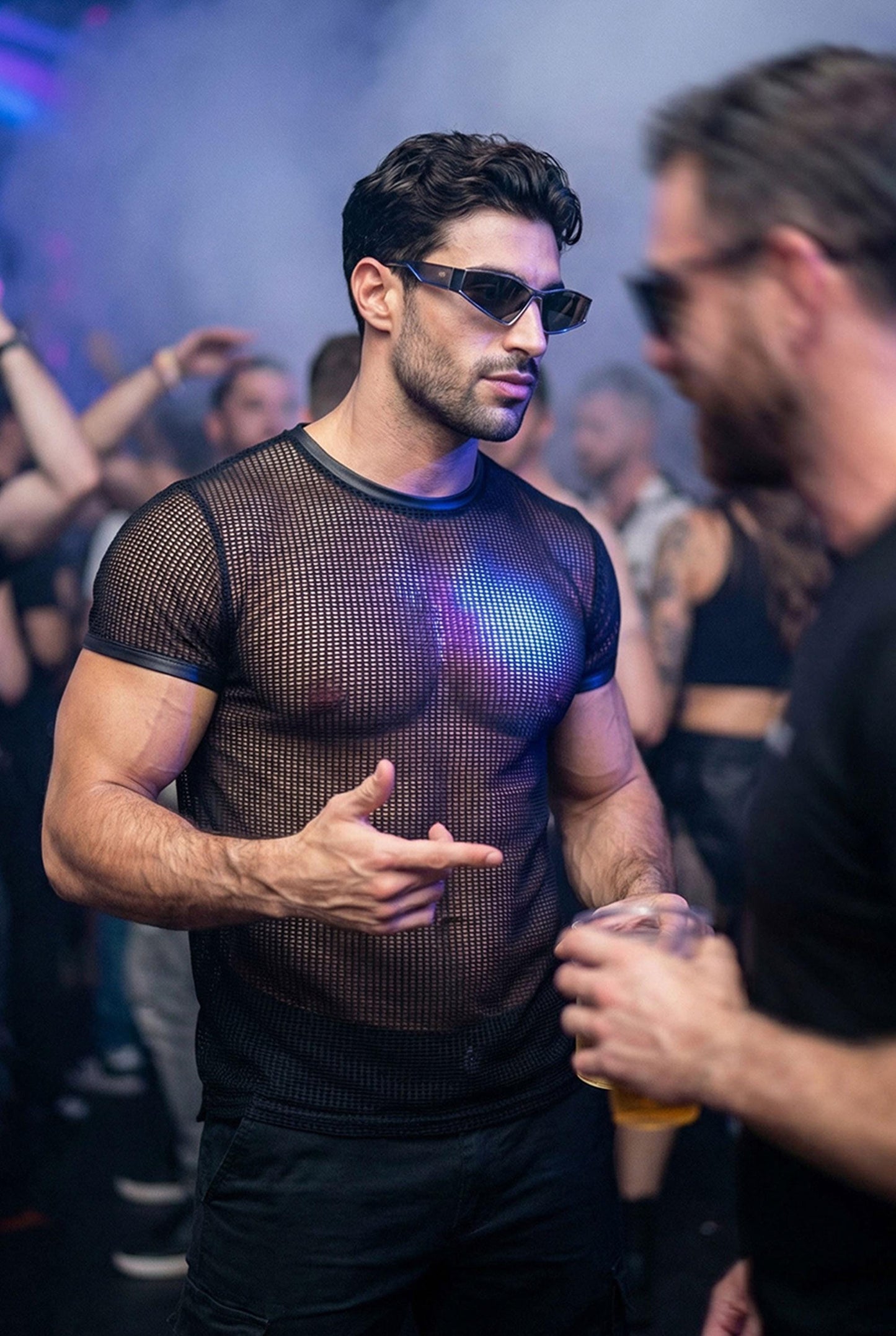 Muscular man wearing a black PIEL men’s mesh top at a crowded nightclub. The fitted short-sleeve mesh top reveals the torso under club lighting while keeping a clean, structured silhouette. PIEL men’s party top designed for nightlife, club wear, and bold queer fashion looks. Confident masculine style, premium mesh top for night events.