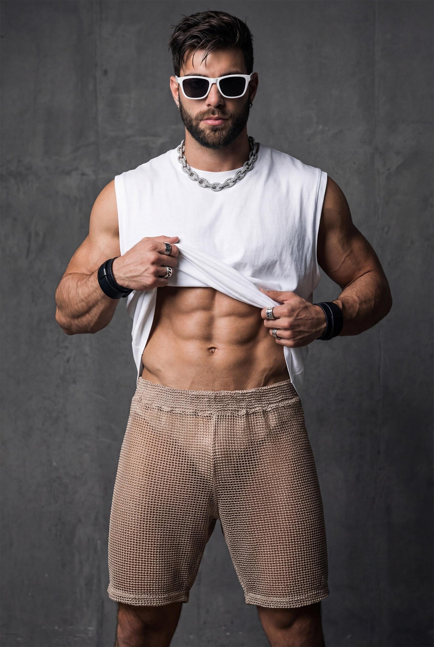 Front view of beige transparent mesh shorts worn by a muscular man. Sheer fabric reveals the body underneath while keeping a fitted, athletic cut. Designed for gay club wear, Berlin nightlife, circuit parties, and pride events. Sexy, breathable party shorts for men who want bold, confident, and eye-catching club outfits.