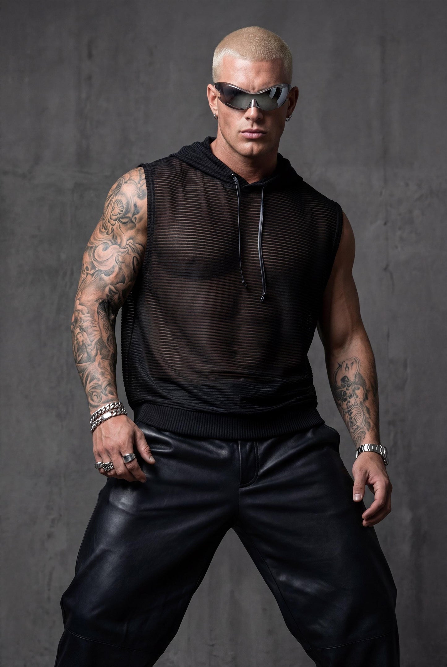 Black mesh hooded tank top men front view showing hood detail and semi-sheer construction with metallic accents. Versatile festival clothing for rave culture enthusiasts, pride parades, and summer adventures. This transparent sleeveless top offers airflow and movement freedom ideal for dancing, performing, or beachside confidence. Bold queer fashion combining urban edge with breathable comfort for nightlife and daytime style.