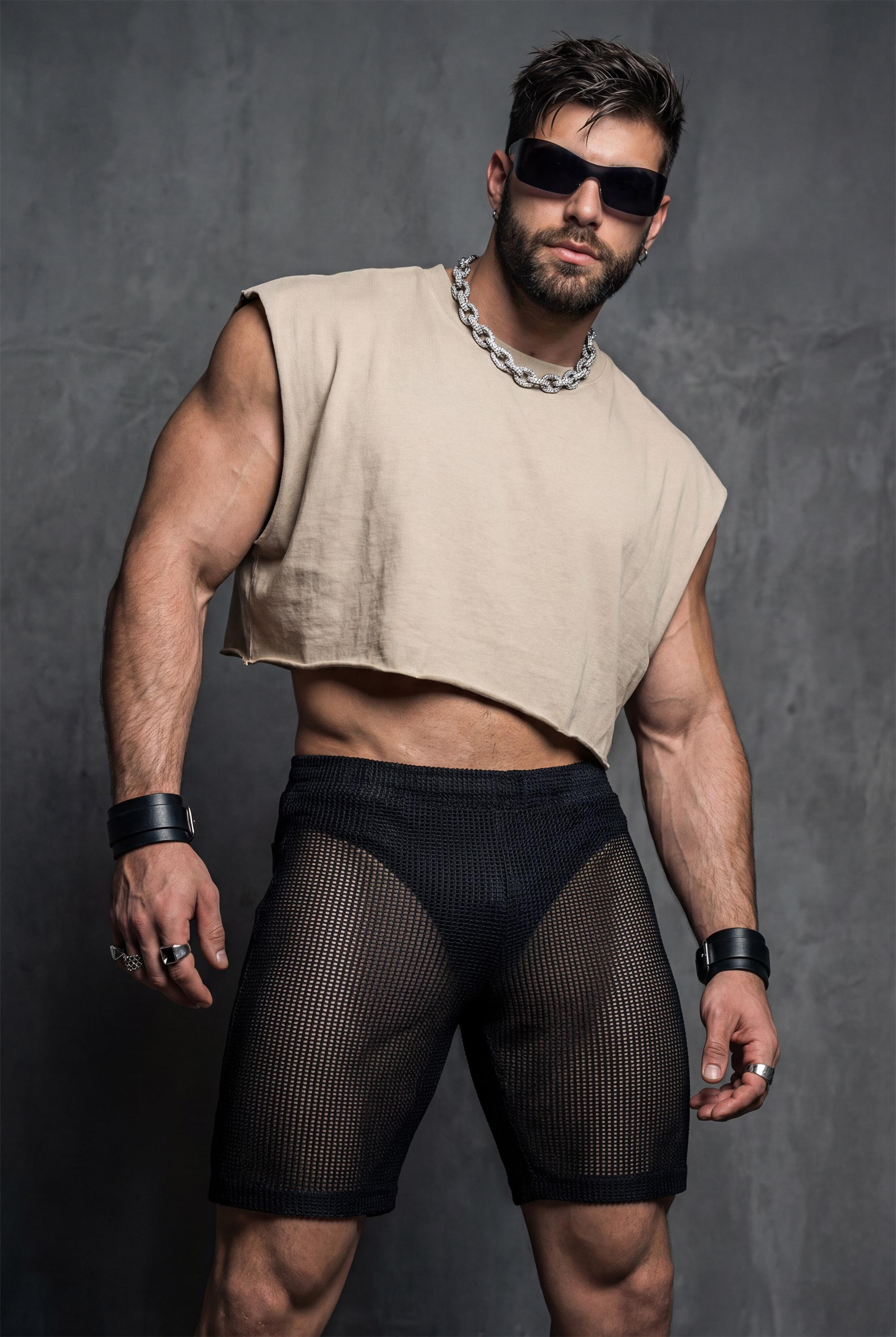 Front view of black transparent mesh shorts worn by a muscular man. Sheer fabric reveals the body underneath while keeping a fitted, athletic cut. Designed for gay club wear, Berlin nightlife, circuit parties, and pride events. Sexy, breathable party shorts for men who want bold, confident, and eye-catching club outfits.