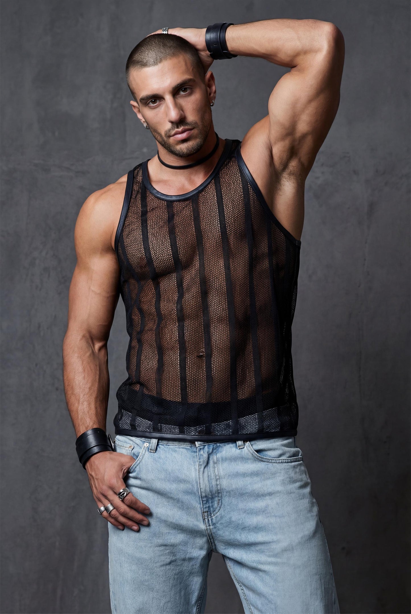 Muscular man styled casually in a black see-through mesh tank top paired with light blue jeans. Transparent racerback stringer with breathable polyester mesh and luxe collar. Versatile men’s party top transitioning from daytime terraces to night clubs, perfect for pride, festivals and statement outfits.