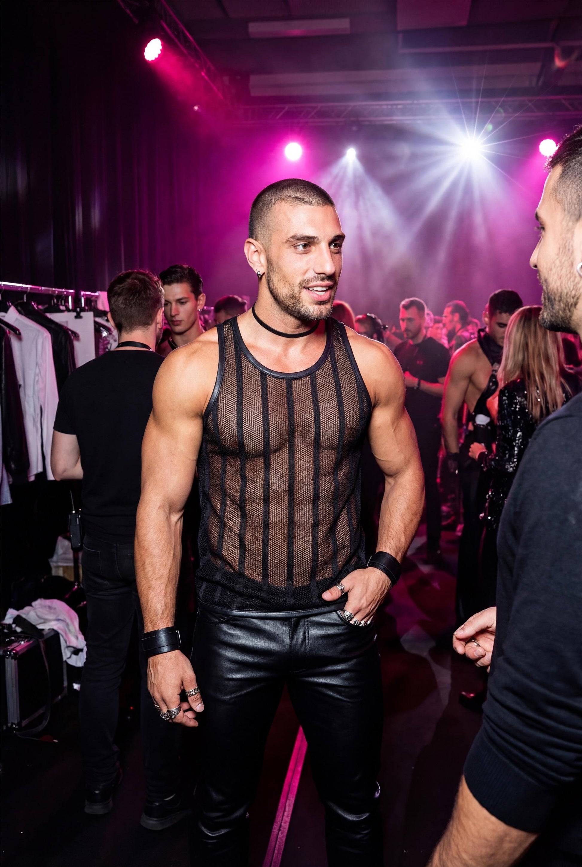 Man wearing a black transparent mesh tank top in a crowded club environment. Racerback stringer design and see-through mesh create a bold party look under club lights. Men’s festival and club wear ideal for circuit parties, pride events, nightlife outfits and gay party fashion.