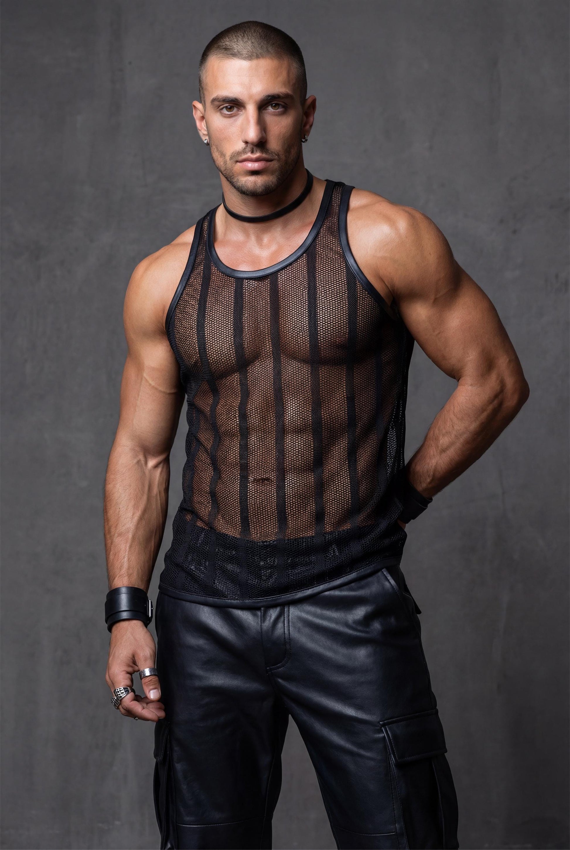 Front view of a muscular man wearing a black transparent mesh tank top. Sleeveless racerback stringer with breathable see-through fabric and clean neckline. Men’s party tank top for club nights, festivals, circuit events, and bold summer nightlife outfits.