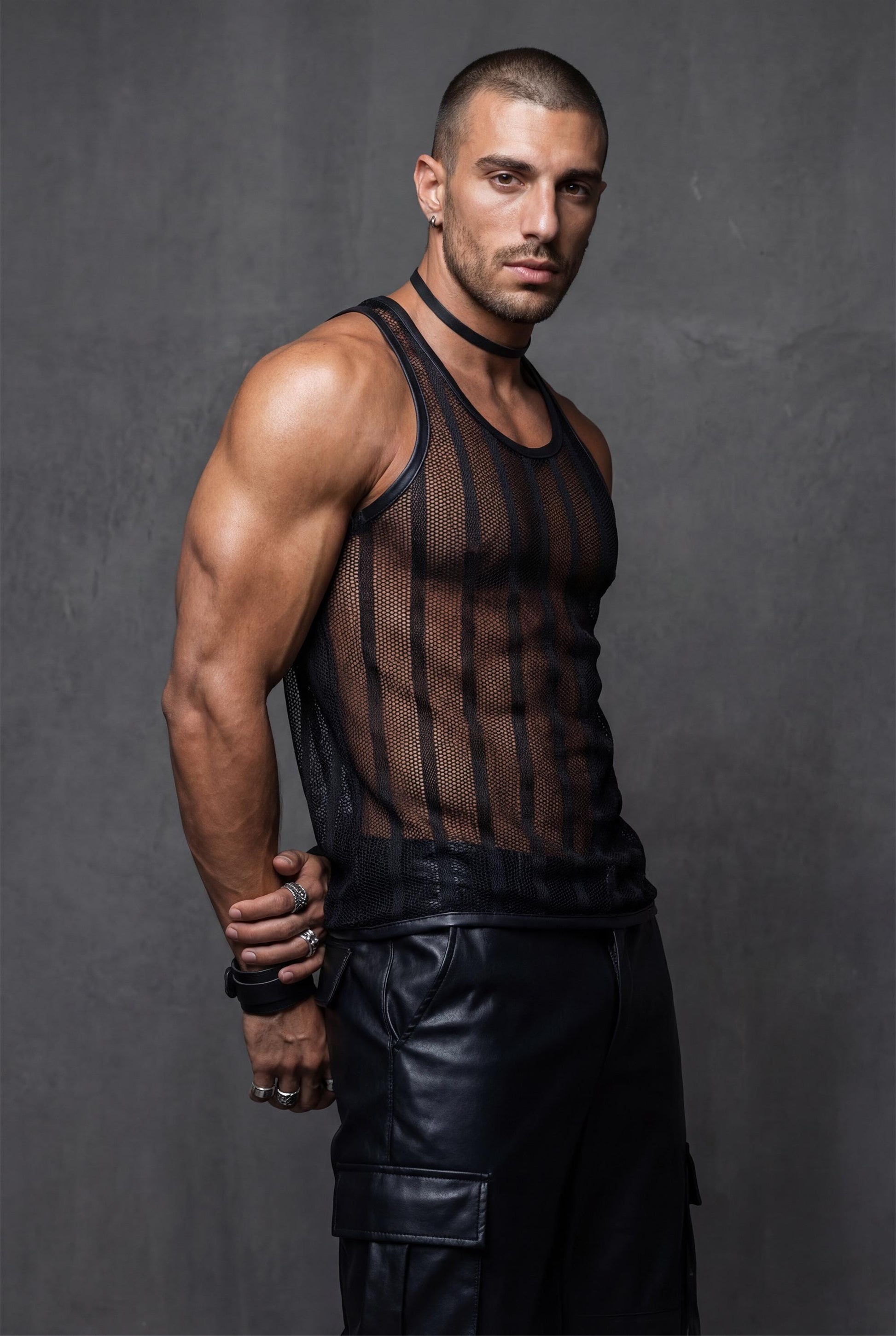Muscular man wearing a black transparent mesh tank top for men, side profile view. See-through racerback stringer reveals chest and abs through breathable polyester mesh, with luxe polyamide collar. Black club party wear styled with leather pants, ideal for gay club nights, pride events, circuit parties, rave festivals and bold men fashion.