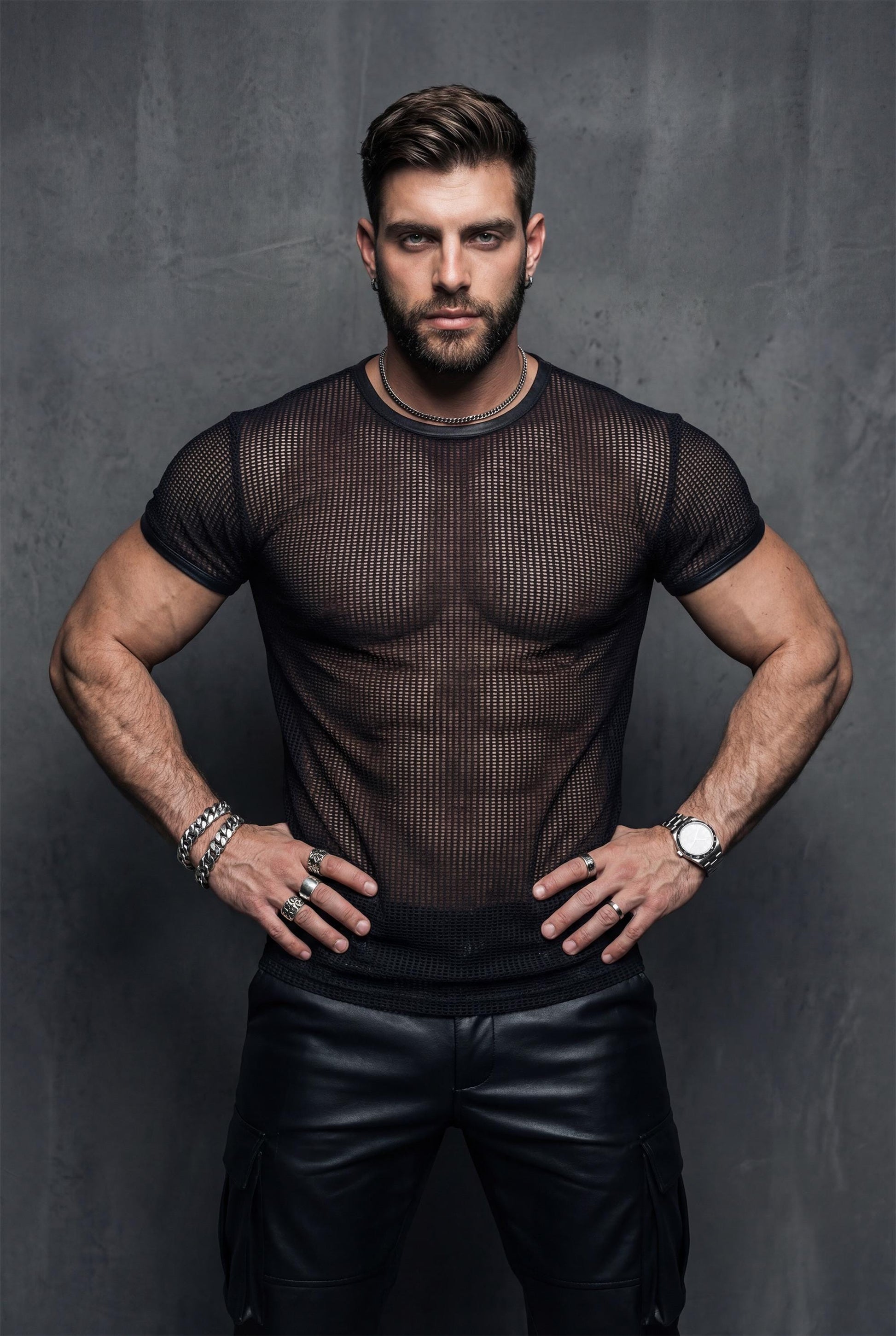 Front view of a muscular man wearing a black see-through mesh t-shirt and leather pants, hands gripping the waistband. The transparent fabric reveals torso definition. Editorial lighting. Men’s mesh top, gay clubwear, fetish fashion, queer nightlife outfit.