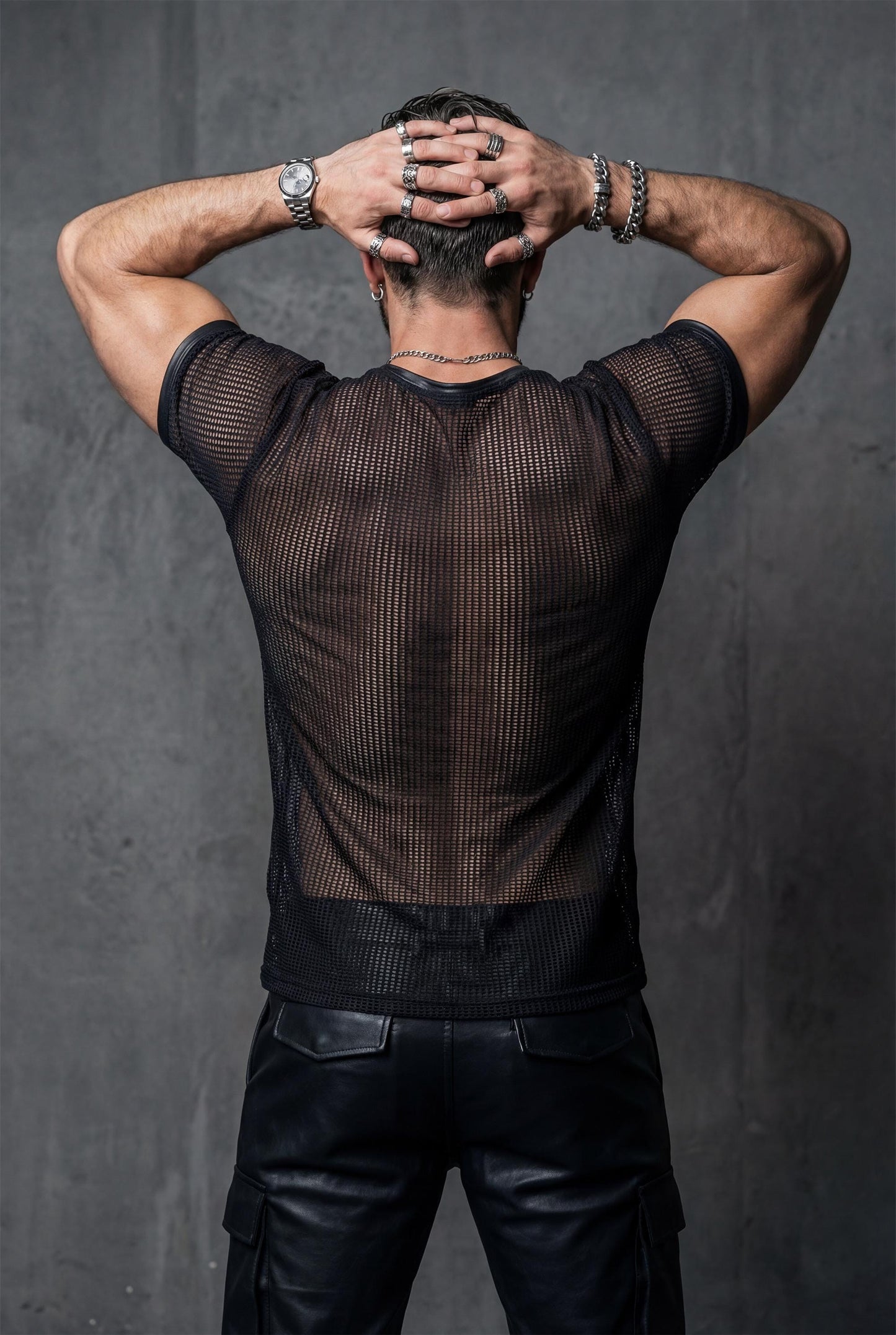 Back view of a muscular man wearing a black transparent mesh t-shirt, arms raised with hands on head. The sheer mesh reveals back muscles and shoulder definition. Dark studio background. Men’s see-through shirt, fetish fashion, gay pride outfit, bold queer clubwear.