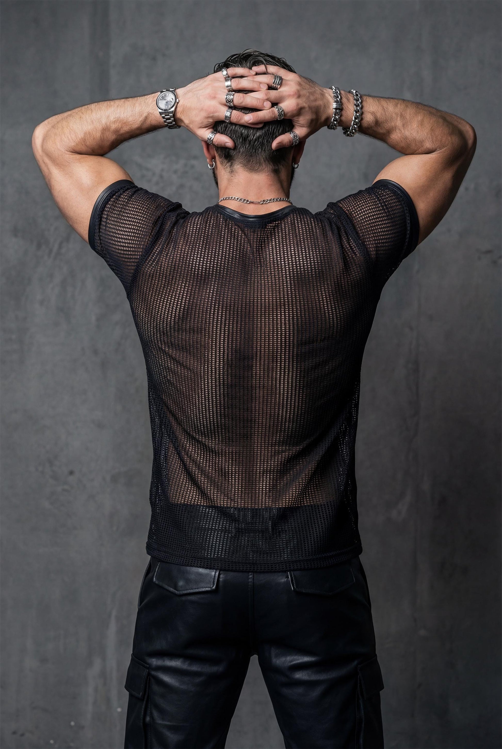 Back view of a muscular man wearing a black transparent mesh t-shirt, arms raised with hands on head. The sheer mesh reveals back muscles and shoulder definition. Dark studio background. Men’s see-through shirt, fetish fashion, gay pride outfit, bold queer clubwear.