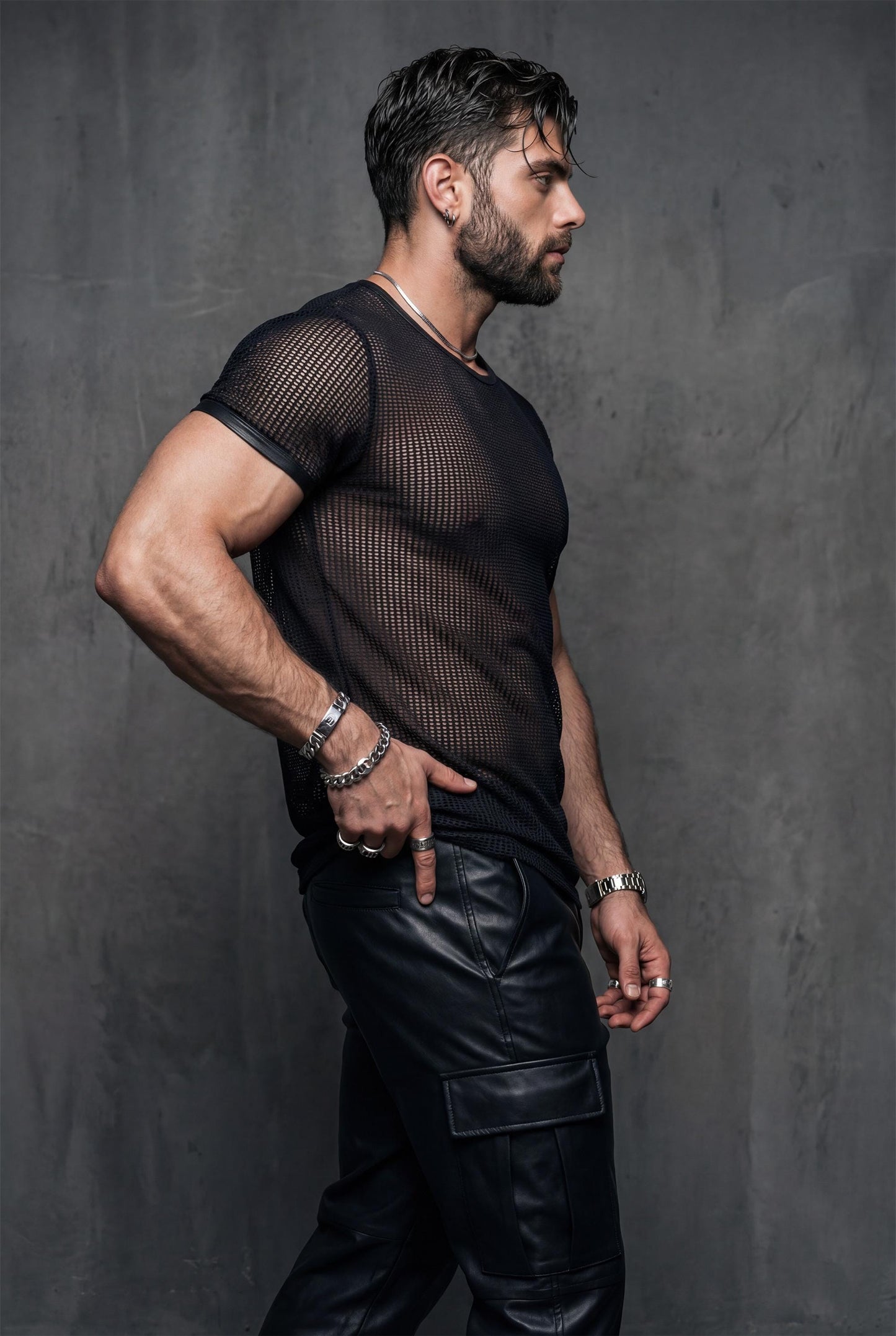 Side profile of a muscular man in a black mesh t-shirt and leather pants, hands behind his back. The transparent fabric outlines chest and arm muscles. Editorial studio lighting, dark background. Men’s sheer mesh shirt, gay clubwear, fetish fashion, masculine queer style.