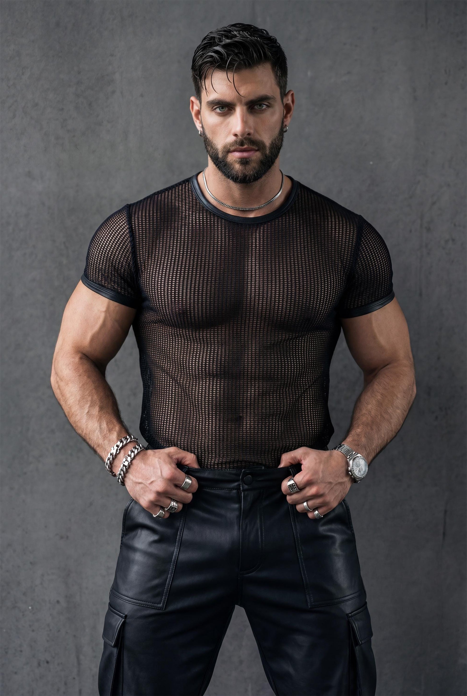 Front view of a muscular man in a black see-through mesh t-shirt and leather pants, hands on hips. The transparent fabric reveals abs and chest with a strong masculine stance. Studio editorial lighting. Men’s mesh top, gay fetish clothing, queer fashion, clubwear and rave outfit.