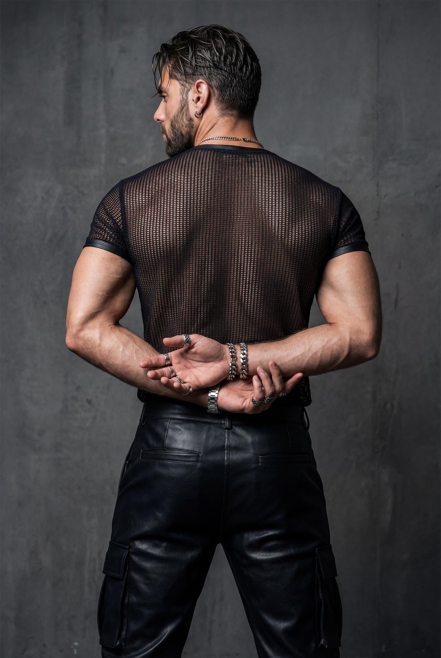 Back view of a muscular man in a black transparent mesh t-shirt, hands crossed behind his back. The sheer fabric reveals the back muscles and spine line. Dark studio background, moody lighting. Men’s see-through shirt, fetish fashion, gay club outfit, queer nightlife clothing, bold masculine style.