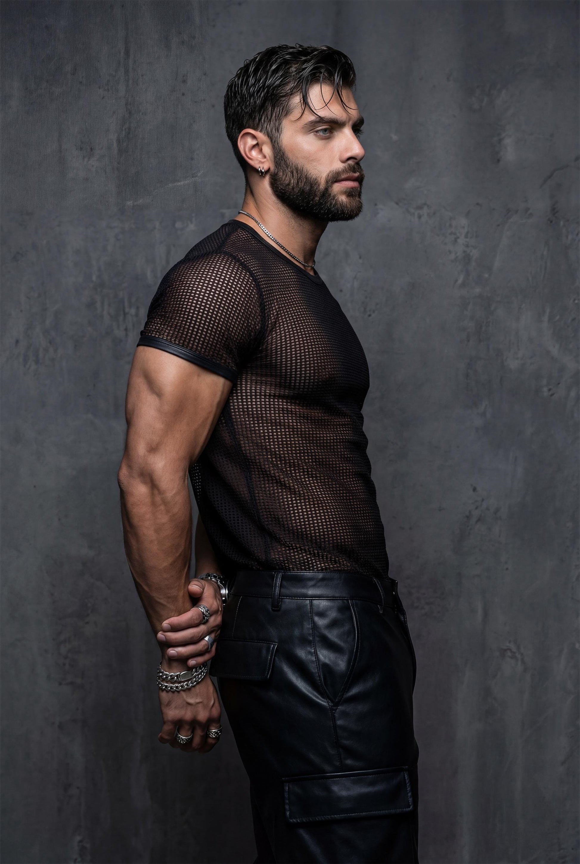 Side profile of a muscular man in a black mesh t-shirt and leather pants, hands behind his back. The transparent fabric outlines chest and arm muscles. Editorial studio lighting, dark background. Men’s sheer mesh shirt, gay clubwear, fetish fashion, masculine queer style.