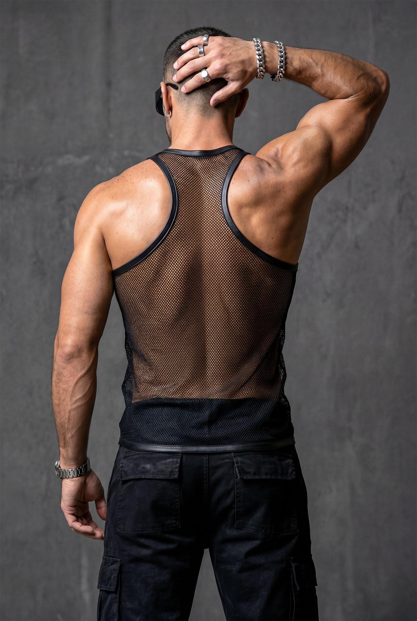 Man wearing a black sheer mesh tank top with faux leather trim around the neckline and armholes. The transparent, breathable fabric highlights the body contours, creating a bold, modern clubwear look. Styled with relaxed blue jeans, this outfit blends sexy confidence with casual streetwear energy