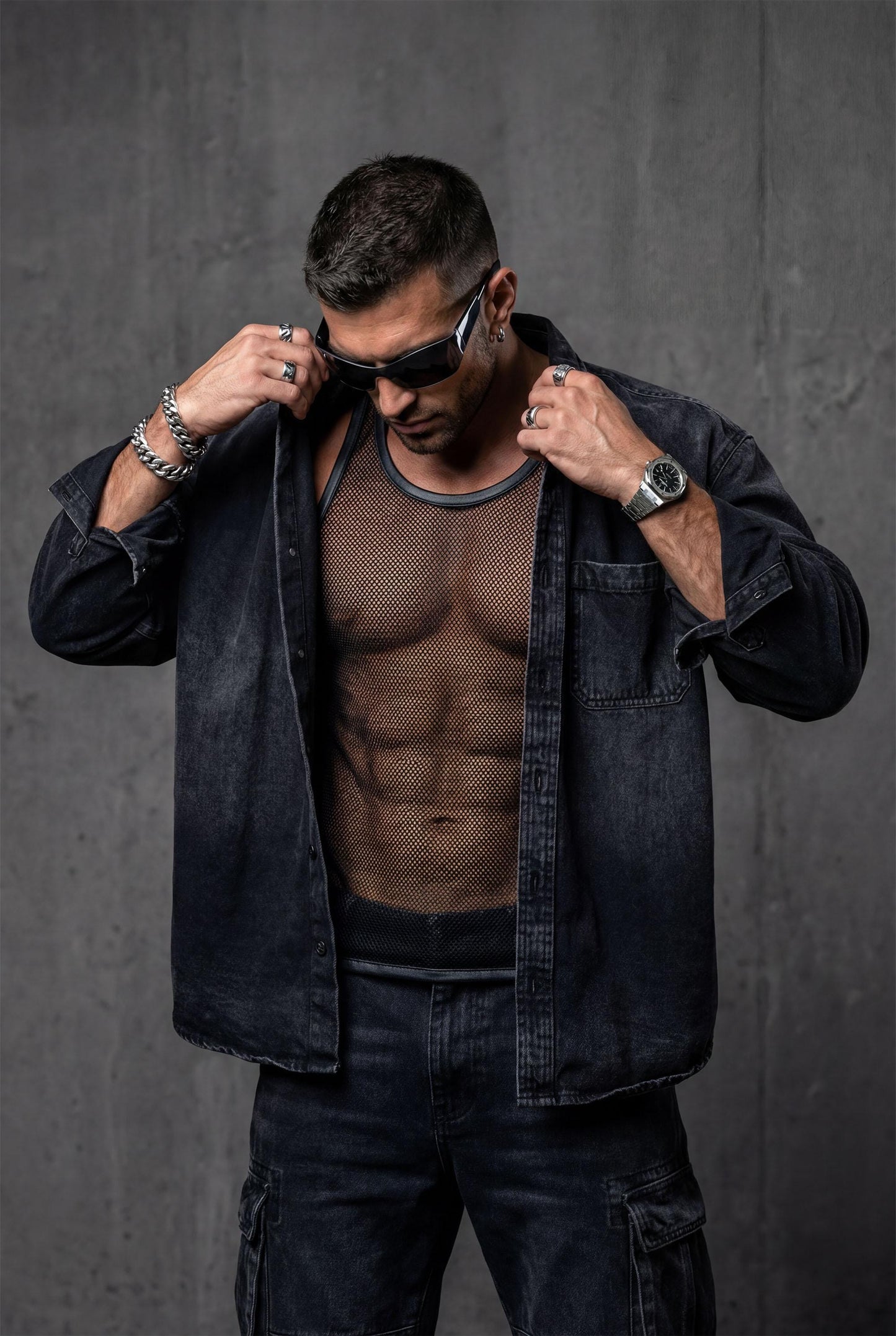Man wearing a black sheer mesh tank top with faux leather trim around the neckline and armholes. The transparent, breathable fabric highlights the body contours, creating a bold, modern clubwear look. Styled with relaxed blue jeans, this outfit blends sexy confidence with casual streetwear energy