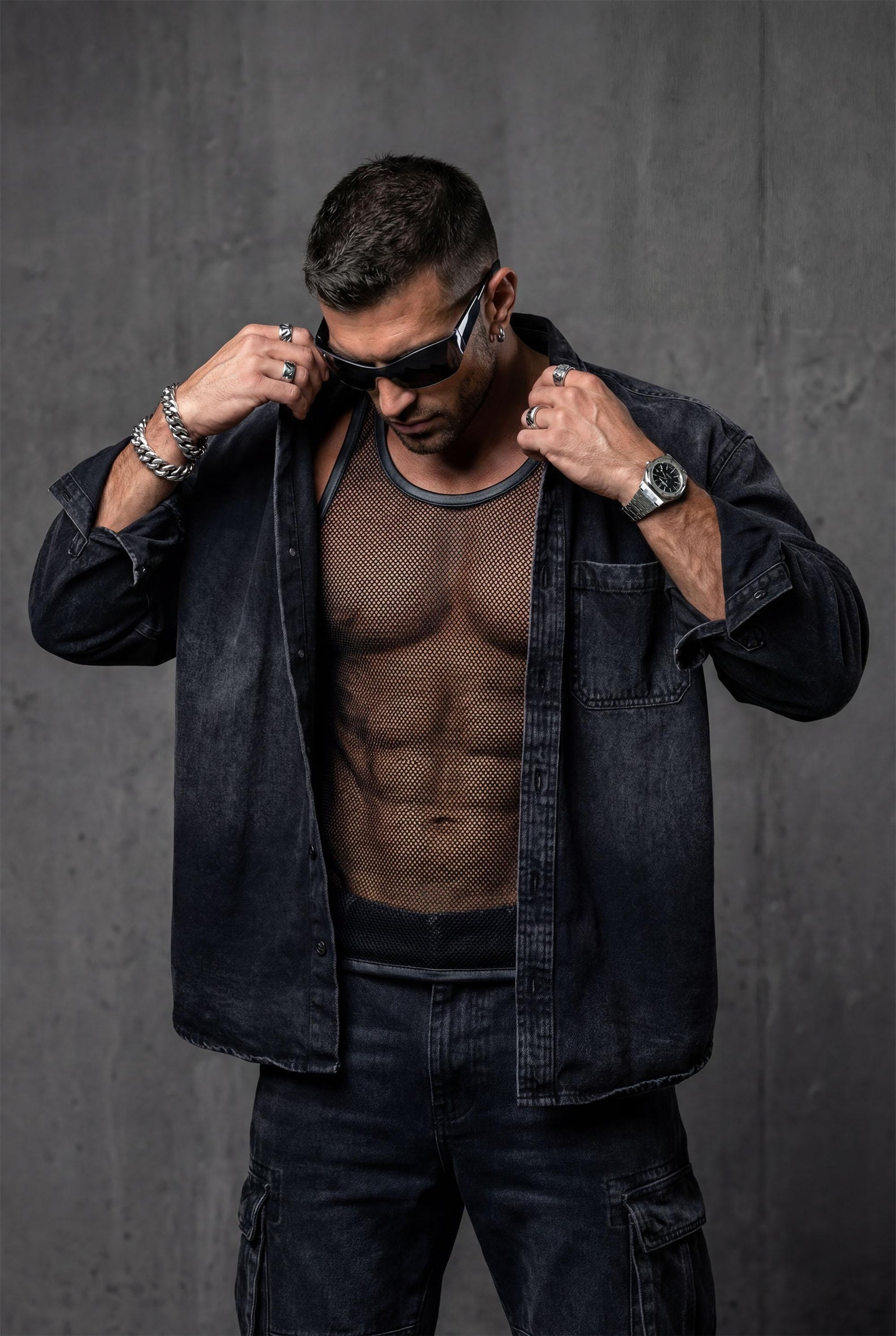 Man wearing a black sheer mesh tank top with faux leather trim around the neckline and armholes. The transparent, breathable fabric highlights the body contours, creating a bold, modern clubwear look. Styled with relaxed blue jeans, this outfit blends sexy confidence with casual streetwear energy