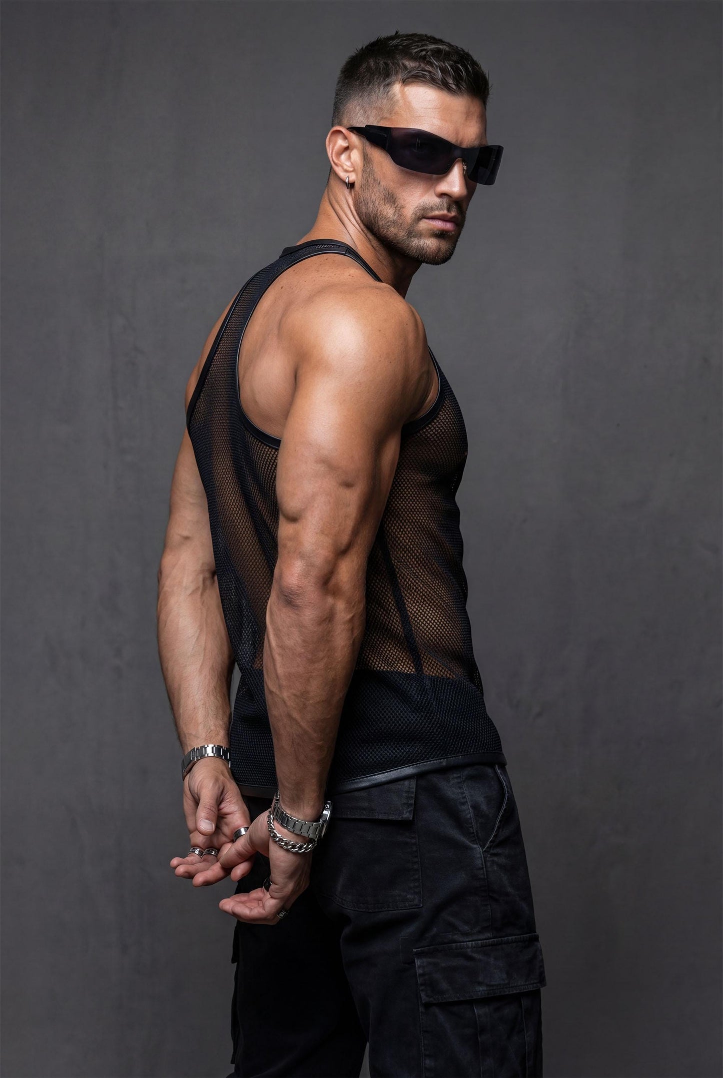 Man wearing a black sheer mesh tank top with faux leather trim around the neckline and armholes. The transparent, breathable fabric highlights the body contours, creating a bold, modern clubwear look. Styled with relaxed blue jeans, this outfit blends sexy confidence with casual streetwear energy