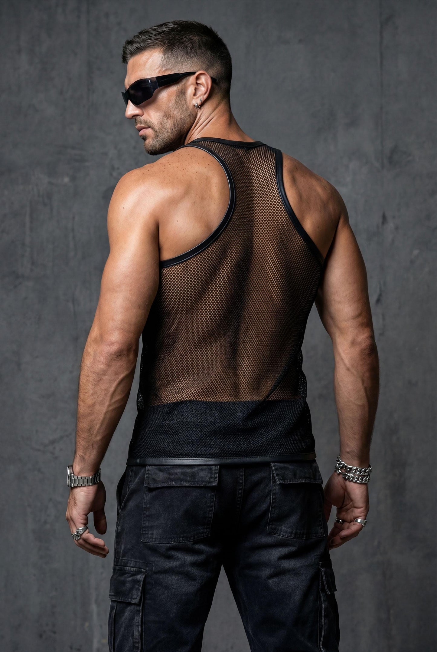 Back view of a man wearing a black sheer mesh tank top with faux leather trim, paired with dark blue jeans. The transparent racerback design reveals the upper back and shoulders, emphasizing the lightweight, breathable fabric — a stylish and confident look for clubwear, nightlife, or summer outfits.