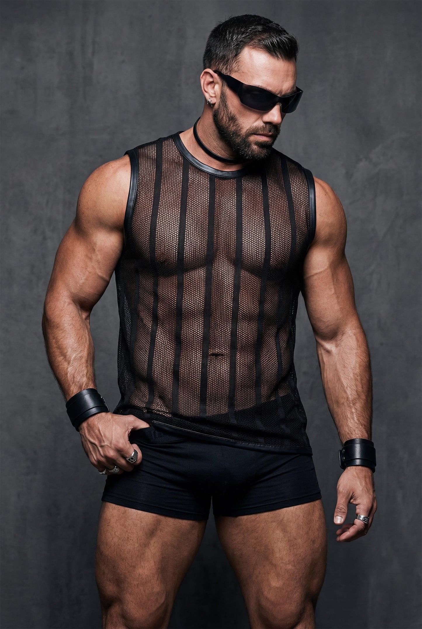 Muscular man wearing a black see-through mesh sleeveless top with vertical stripes, styled with black fitted shorts. The transparent fabric reveals chest and abs. Studio lighting, bold masculine look, gay clubwear and pride festival outfit.