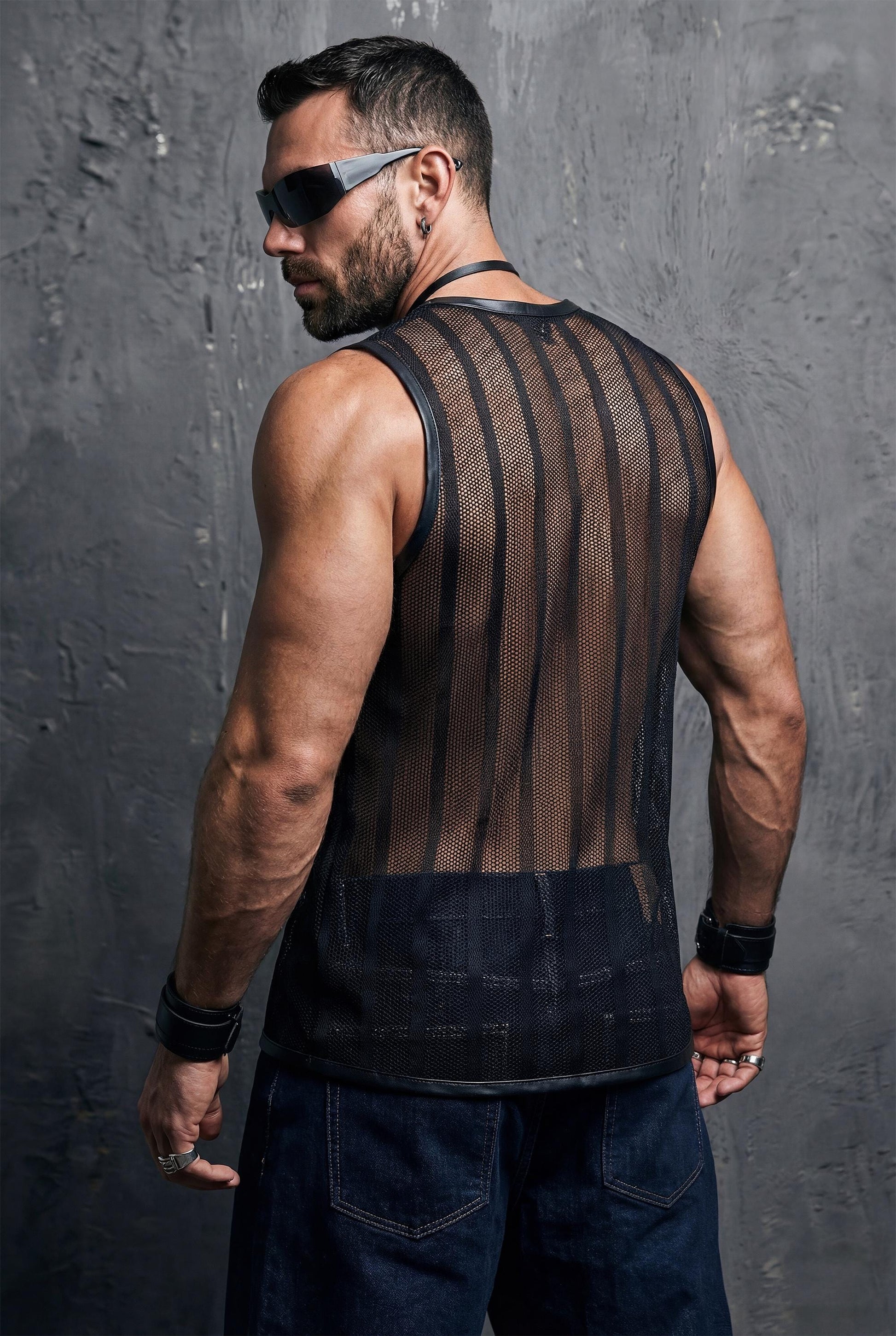 Muscular man wearing a black sheer mesh sleeveless top with vertical stripes, photographed from the back. The transparent mesh reveals strong shoulders and back muscles, finished with vegan leather trim. Dark studio background, masculine clubwear look, gay festival and rave fashion style.