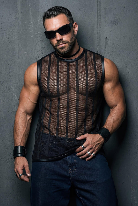 Muscular man leaning against a wall wearing a black transparent mesh sleeveless shirt with vertical stripes. Sheer fabric reveals chest and abs, styled with dark jeans. Strong masculine attitude, studio fashion photography, gay clubwear style.