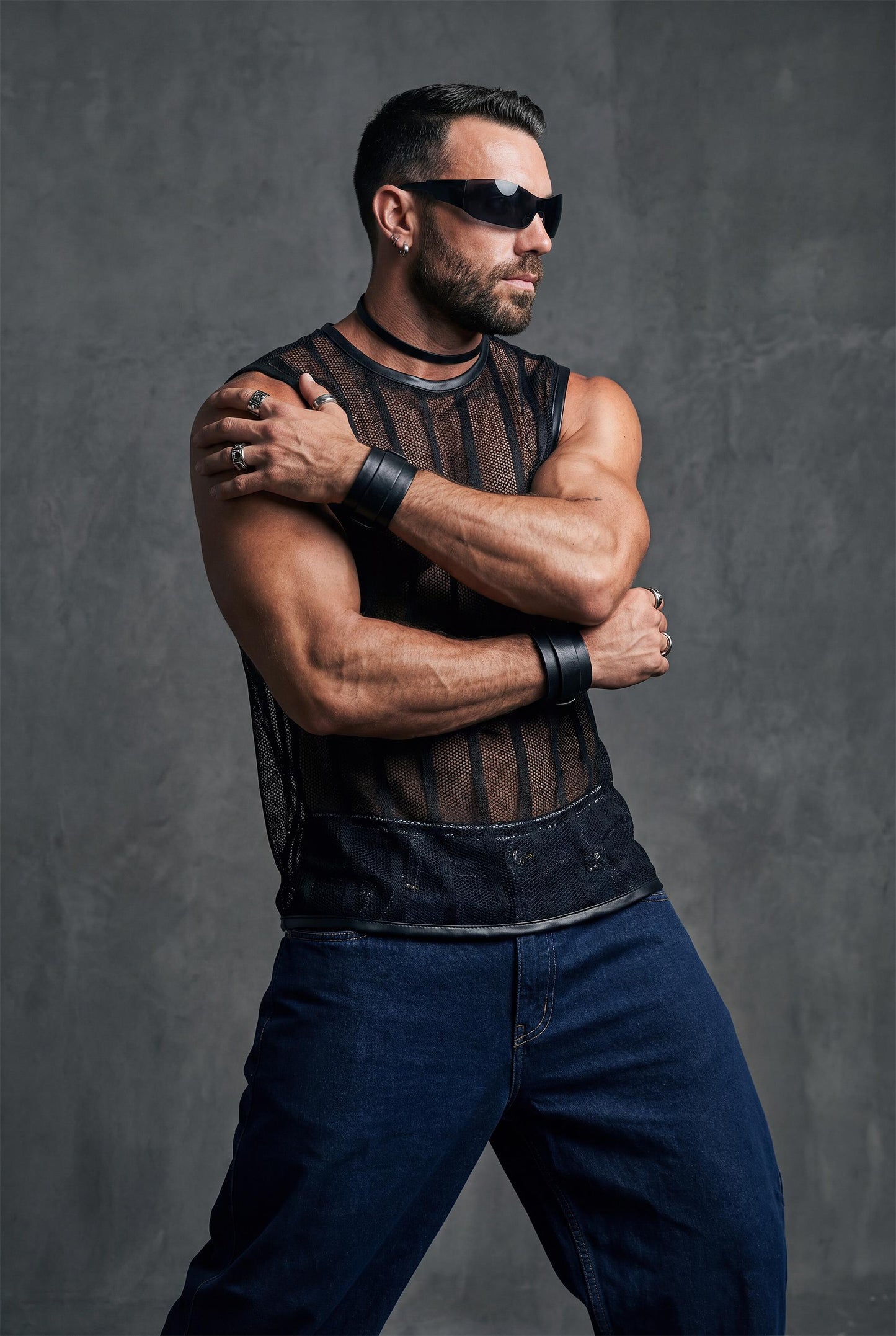 Muscular man posing with crossed arms wearing a black transparent mesh sleeveless shirt with vertical stripes. The sheer fabric reveals the chest and torso, paired with dark jeans. Strong masculine attitude, studio fashion shoot, men’s gay club and festival wear.