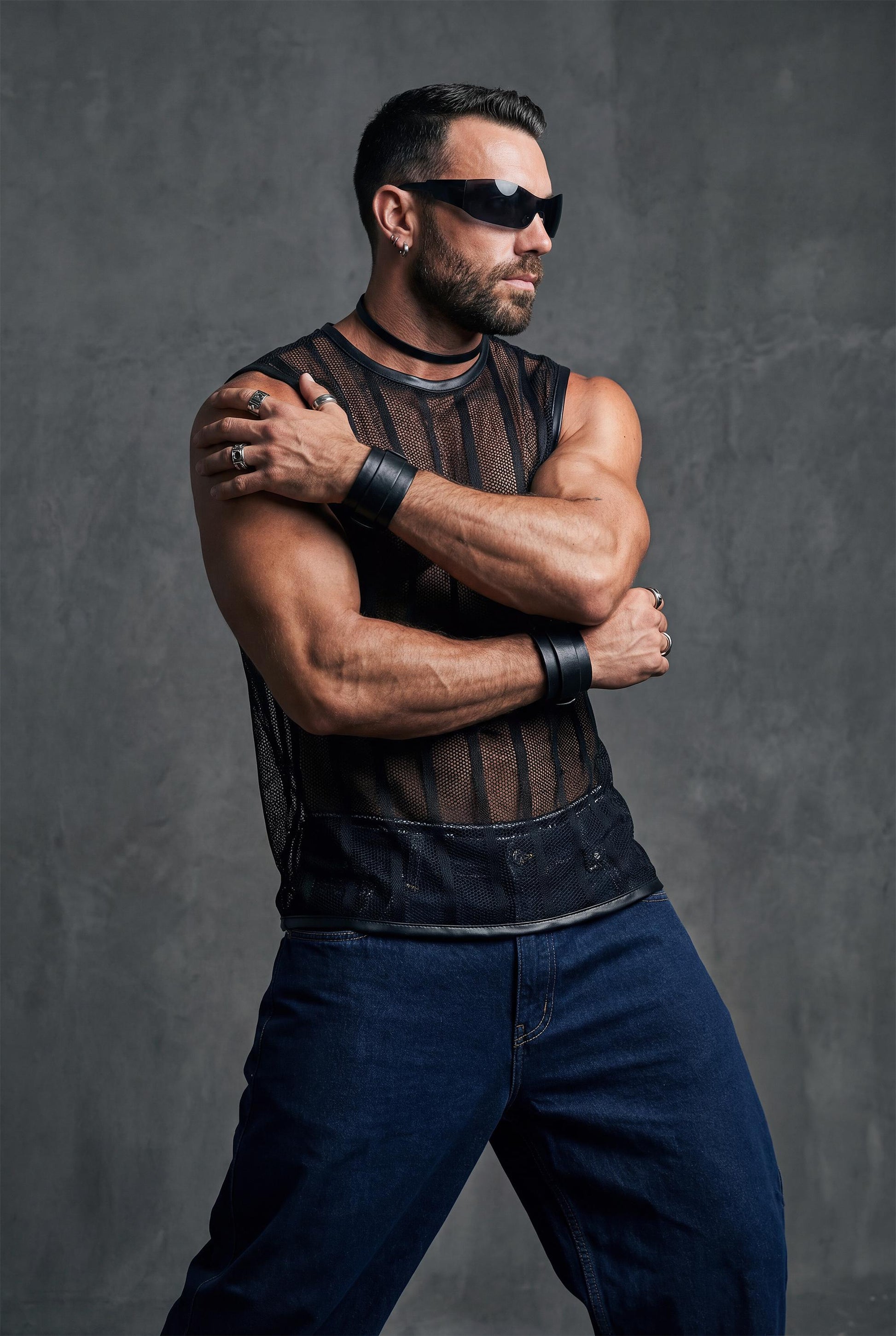 Muscular man posing with crossed arms wearing a black transparent mesh sleeveless shirt with vertical stripes. The sheer fabric reveals the chest and torso, paired with dark jeans. Strong masculine attitude, studio fashion shoot, men’s gay club and festival wear.