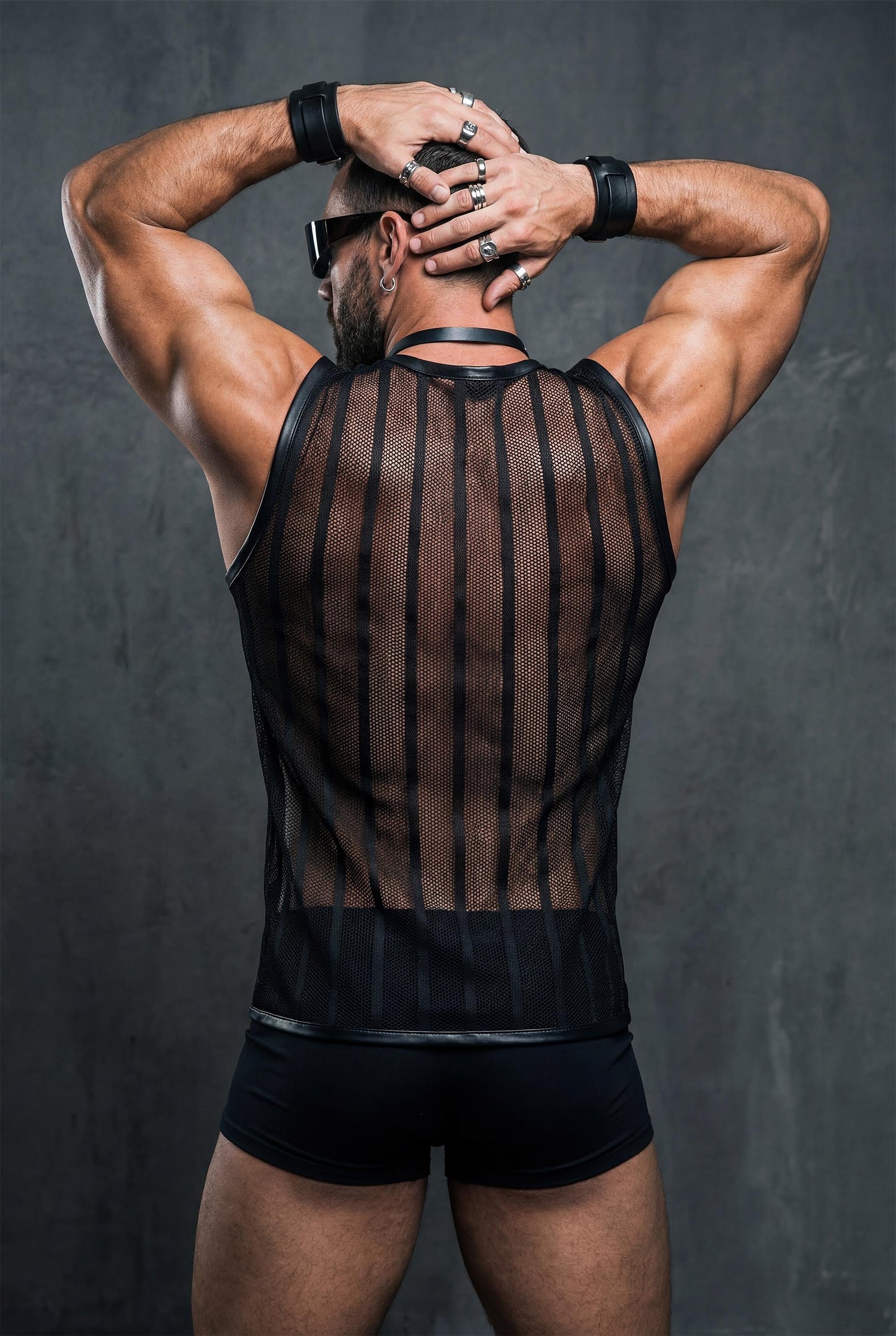 Back view of a muscular man raising his arms while wearing a black transparent mesh tank top. Vertical striped mesh highlights shoulder and back muscles. Strong masculine pose, studio editorial style, men’s rave and club fashion.