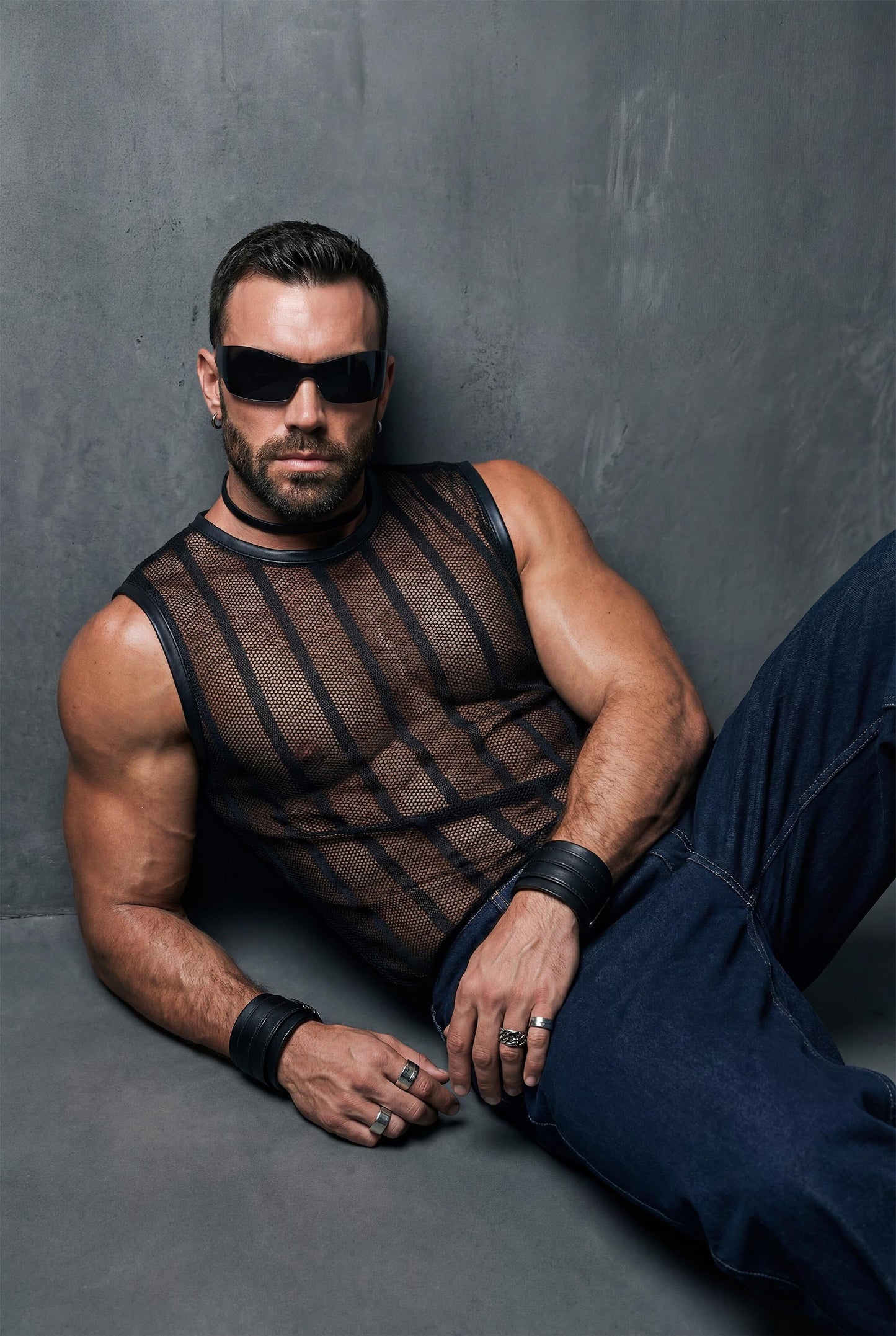 Muscular man reclining on the floor wearing a black sheer mesh sleeveless top with vertical stripes and dark jeans. Transparent fabric reveals torso and chest. Editorial studio shoot, masculine gay fashion, club and nightlife style.