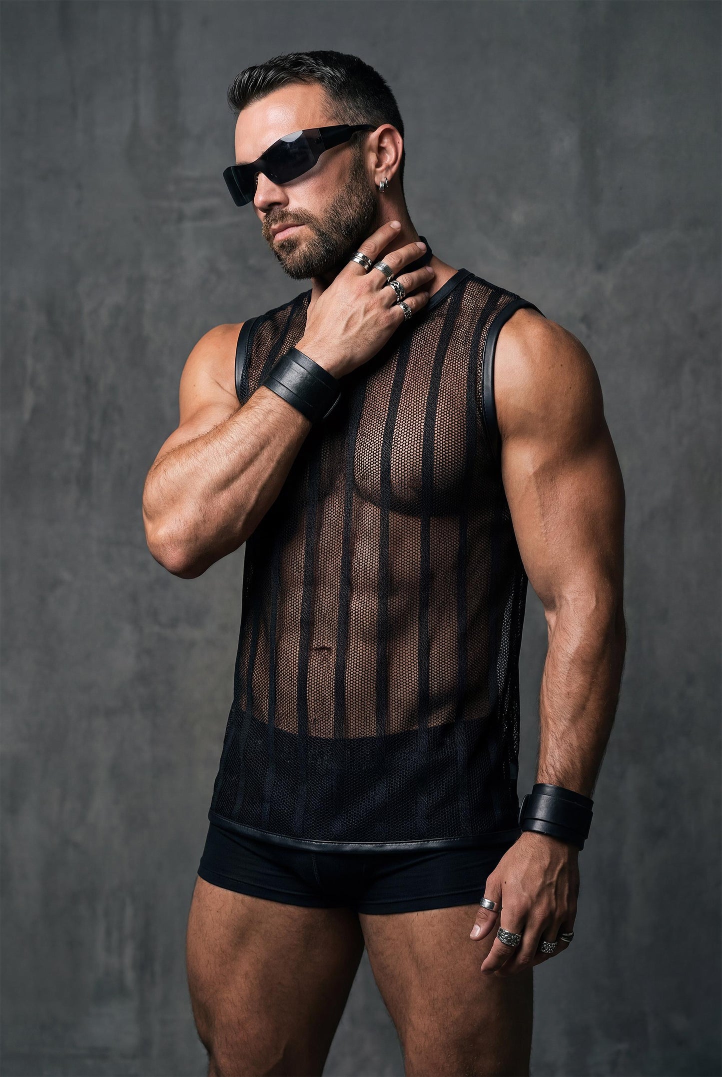 Muscular man wearing a black see-through mesh sleeveless top with vertical stripes, styled with black fitted shorts. The transparent fabric reveals chest and abs. Studio lighting, bold masculine look, gay clubwear and pride festival outfit.