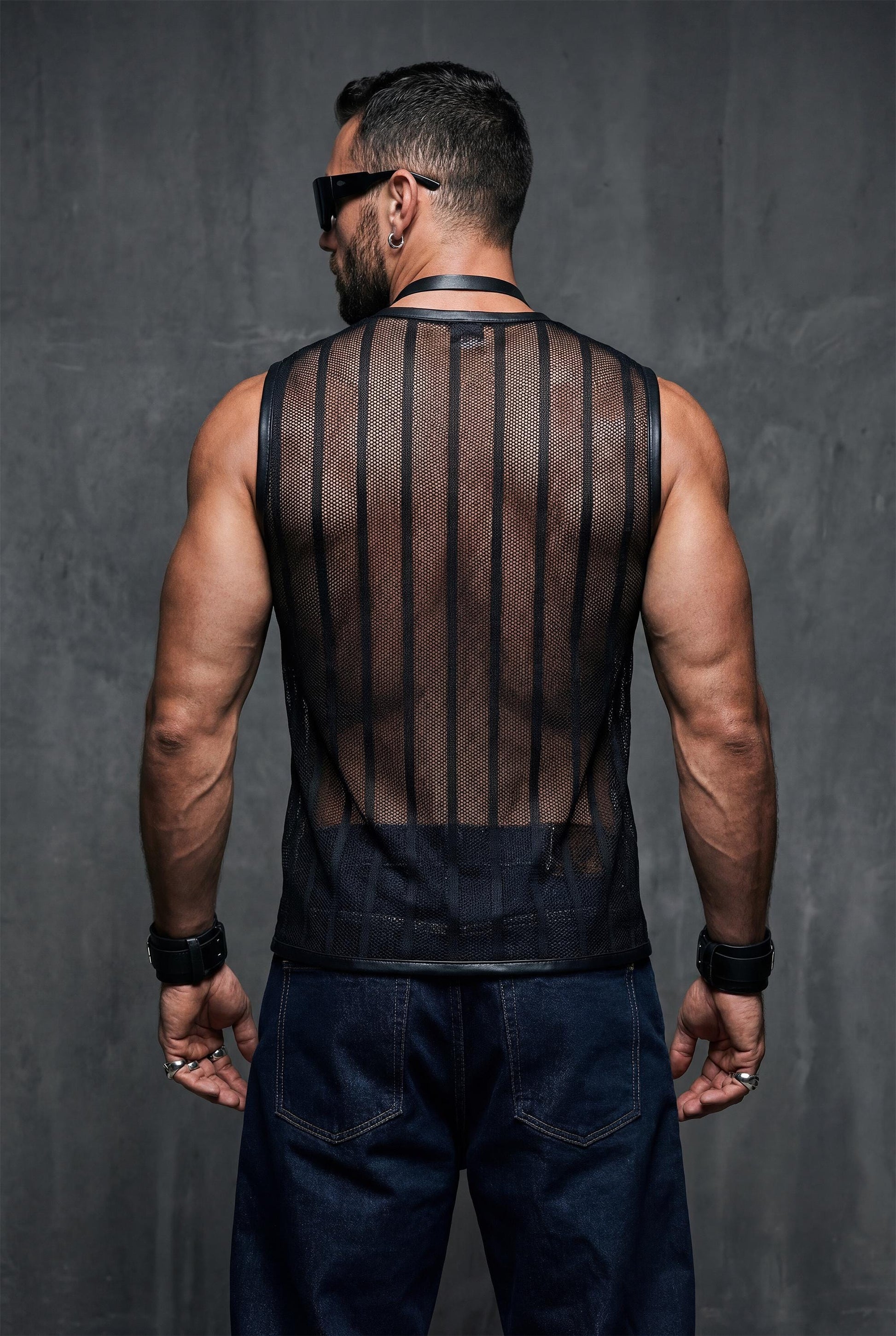 Back view of a muscular man in a black see-through mesh tank top with vertical striped pattern. Transparent fabric highlights the back and arms, finished with black trim. Editorial studio lighting, men’s clubwear, rave outfit, gay pride fashion.