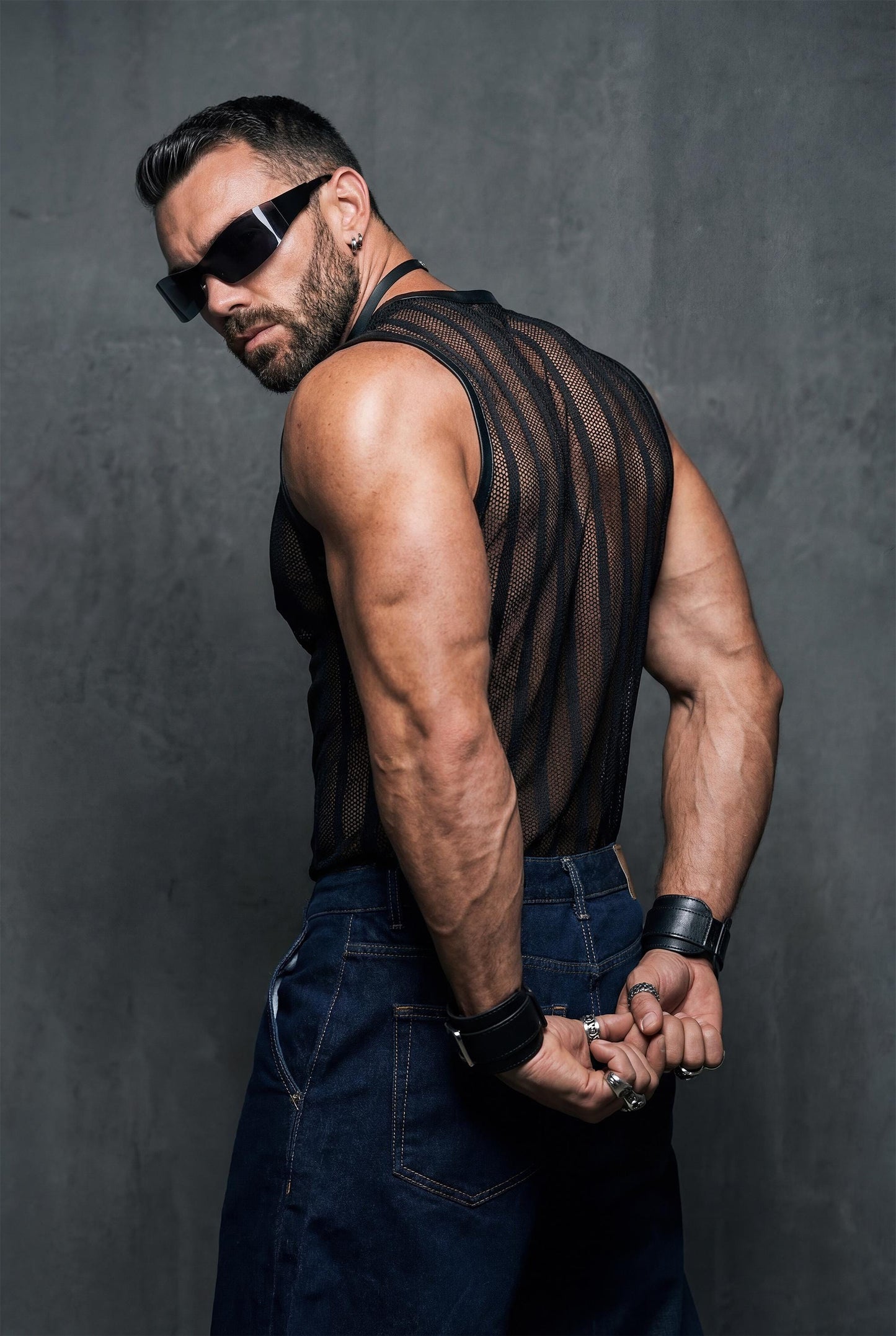 Side back view of a muscular man with hands behind his back, wearing a black see-through mesh tank top. The striped transparent fabric emphasizes arm and back definition. Studio background, bold men’s clubwear, gay fetish-inspired fashion.