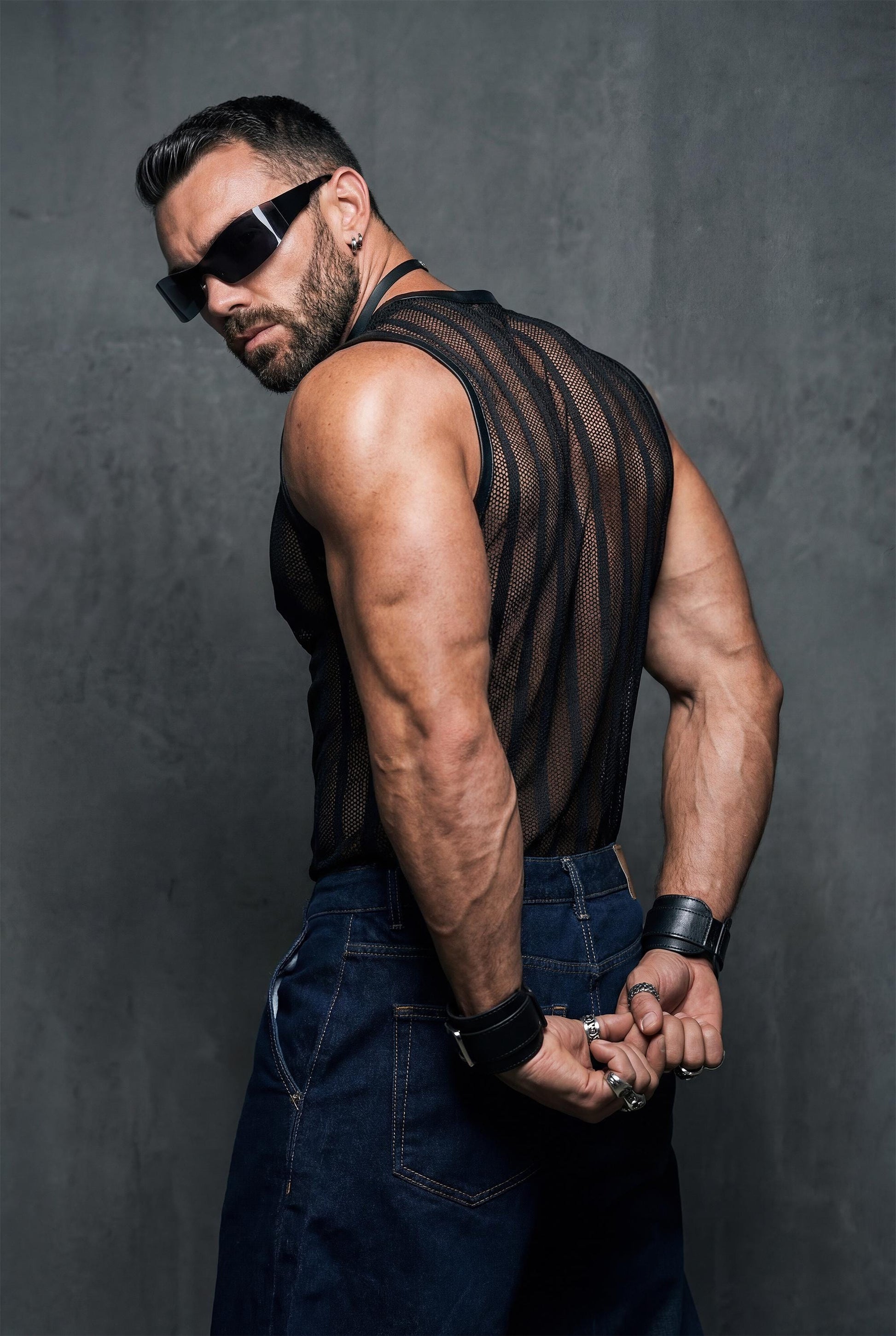 Side back view of a muscular man with hands behind his back, wearing a black see-through mesh tank top. The striped transparent fabric emphasizes arm and back definition. Studio background, bold men’s clubwear, gay fetish-inspired fashion.