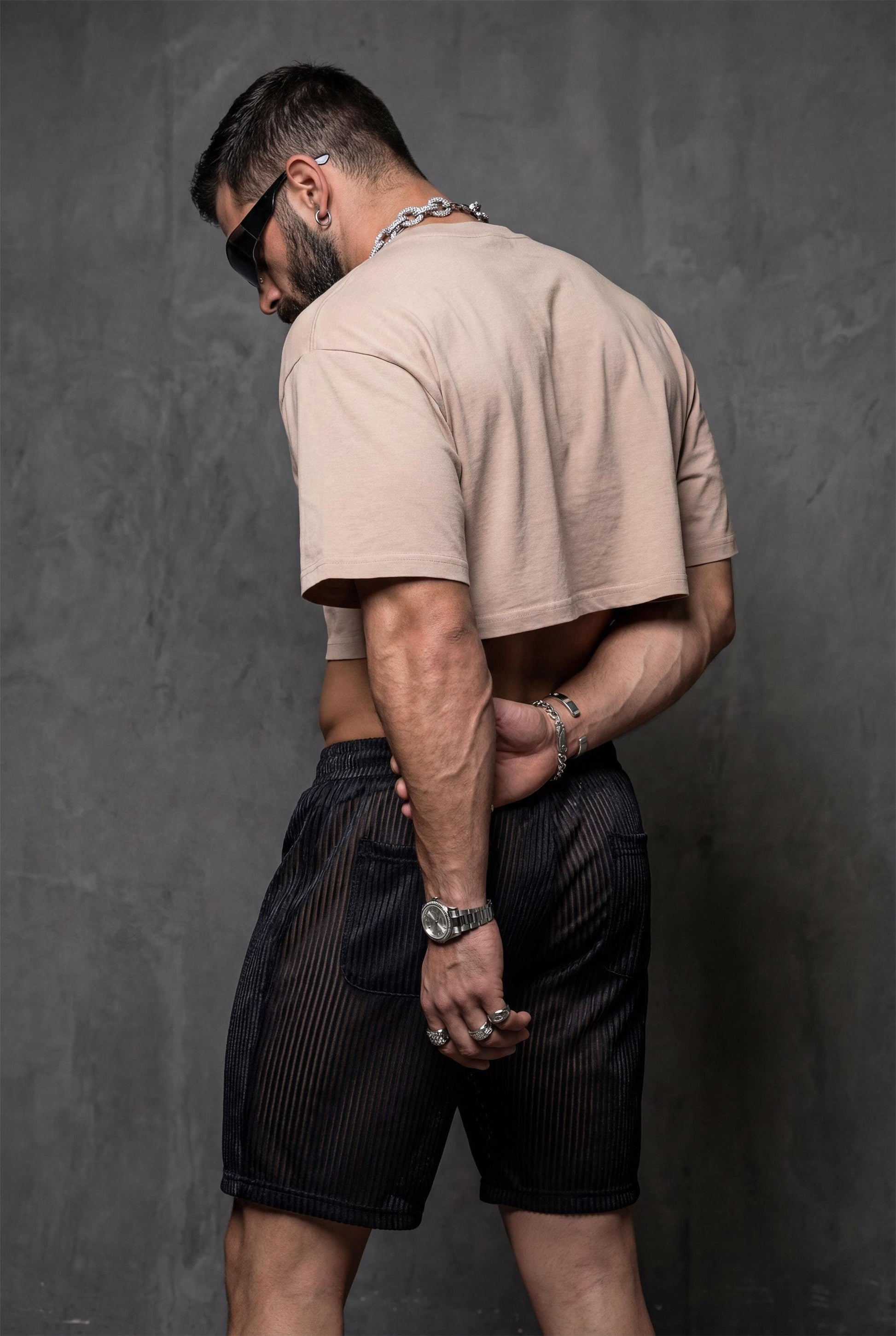 Black semi-transparent mesh shorts men with metallic pinstripe styled with white tank for casual rave outfit and gay festival fashion. Shimmering shorts show versatility paired with different tops. Perfect for circuit parties, pride parades, beach events, and summer celebrations. Statement look for bold wardrobe with confident shimmering expression.