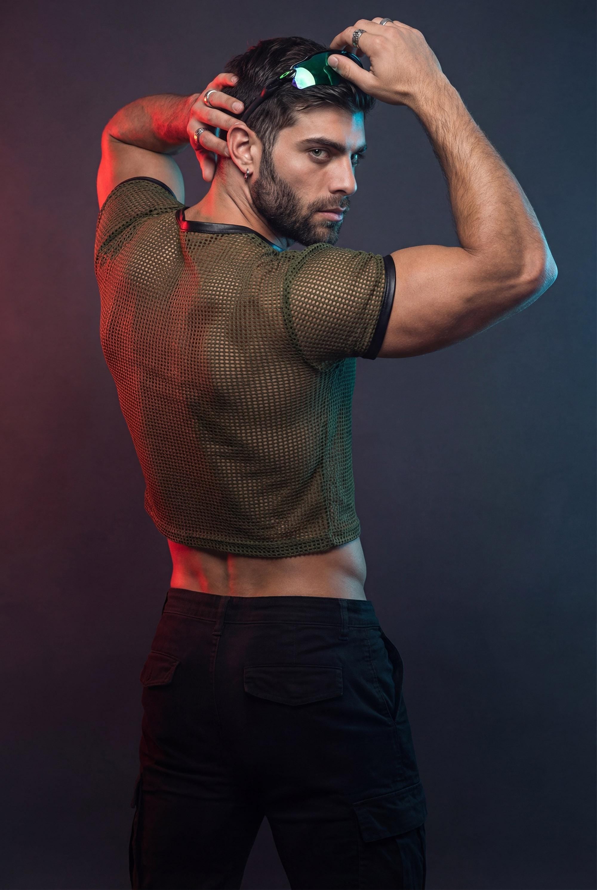 Muscular man wearing an olive green PIEL men’s mesh crop top in a studio setting with red and blue lighting. The short-sleeve mesh top reveals the back and arms while keeping a fitted, structured cut. PIEL men’s party top designed for club wear, nightlife outfits, and bold queer fashion aesthetics. Confident masculine style, premium mesh top.