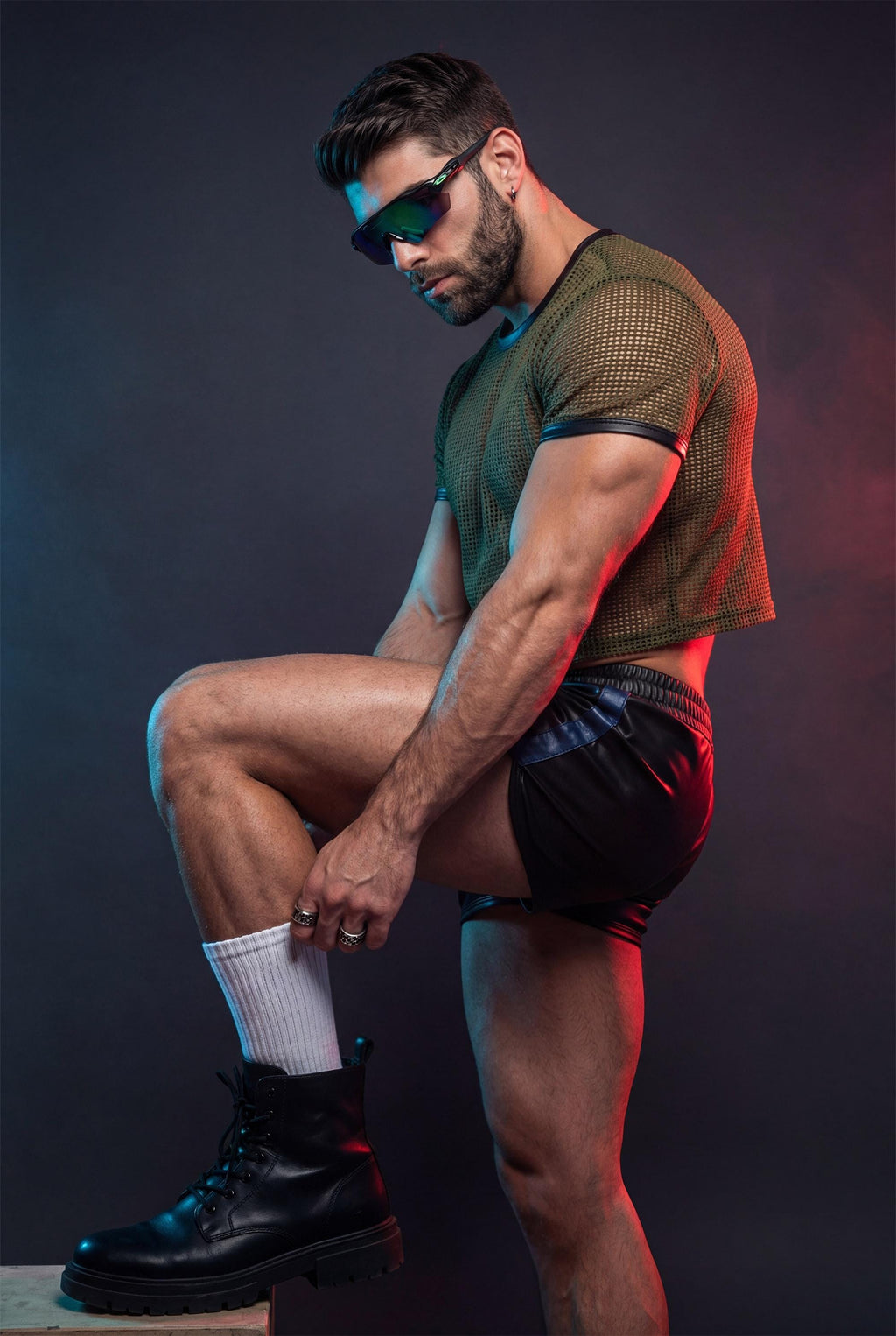 Muscular man wearing an olive green PIEL men’s mesh crop top in a studio setting with red and blue lighting. The short-sleeve mesh top reveals the back and arms while keeping a fitted, structured cut. PIEL men’s party top designed for club wear, nightlife outfits, and bold queer fashion aesthetics. Confident masculine style, premium mesh top.