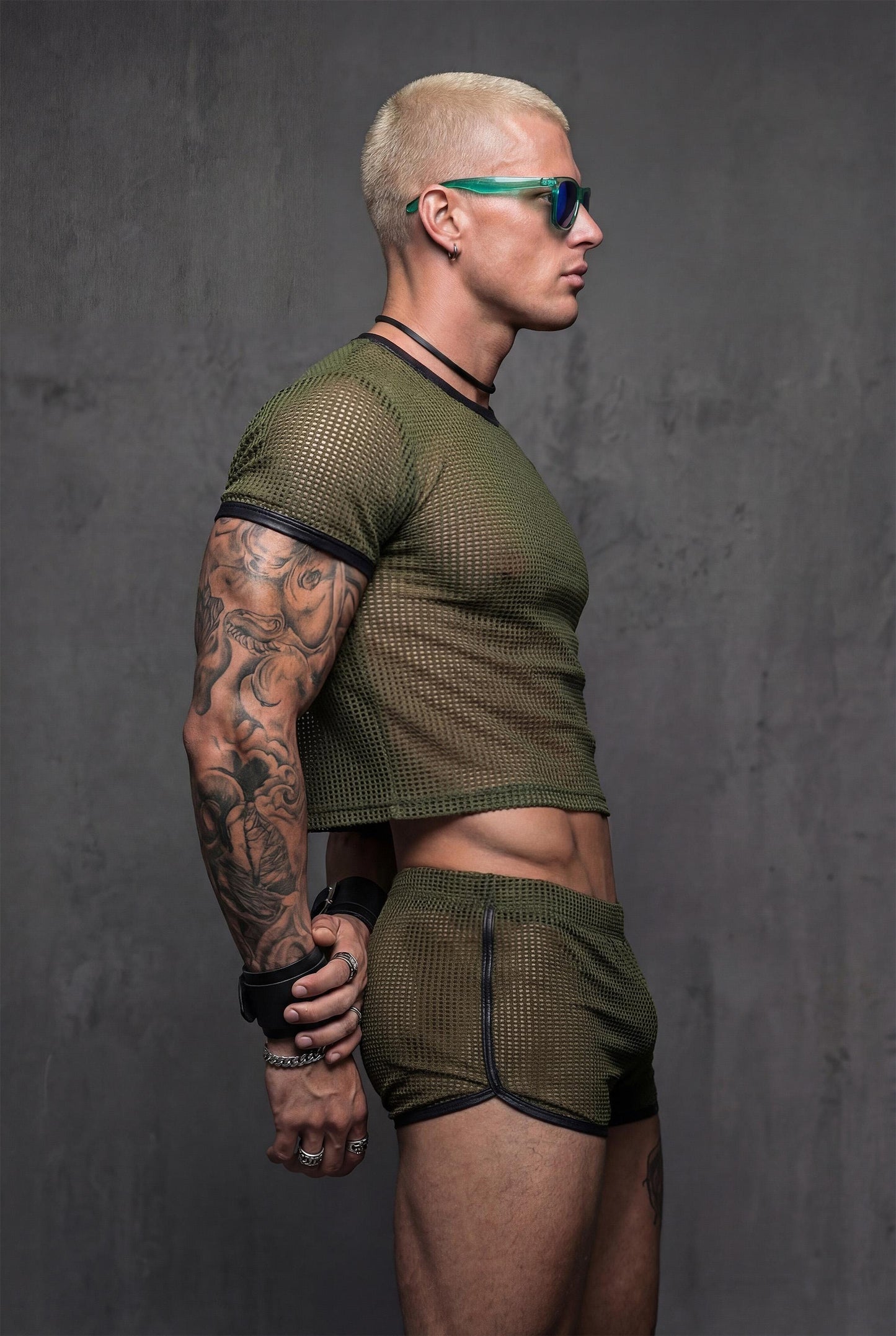 Side profile military green sheer mesh crop top with black vegan leather contrast trim paired with transparent shorts - PIEL Night Starter complete look for gay circuit parties underground raves - athletic body-hugging fit reveals masculine physique with confidence - queer menswear perfect for techno festivals pride parades fetish clubs beach parties - olive green alternative color stands out at rave events lgbtq celebrations