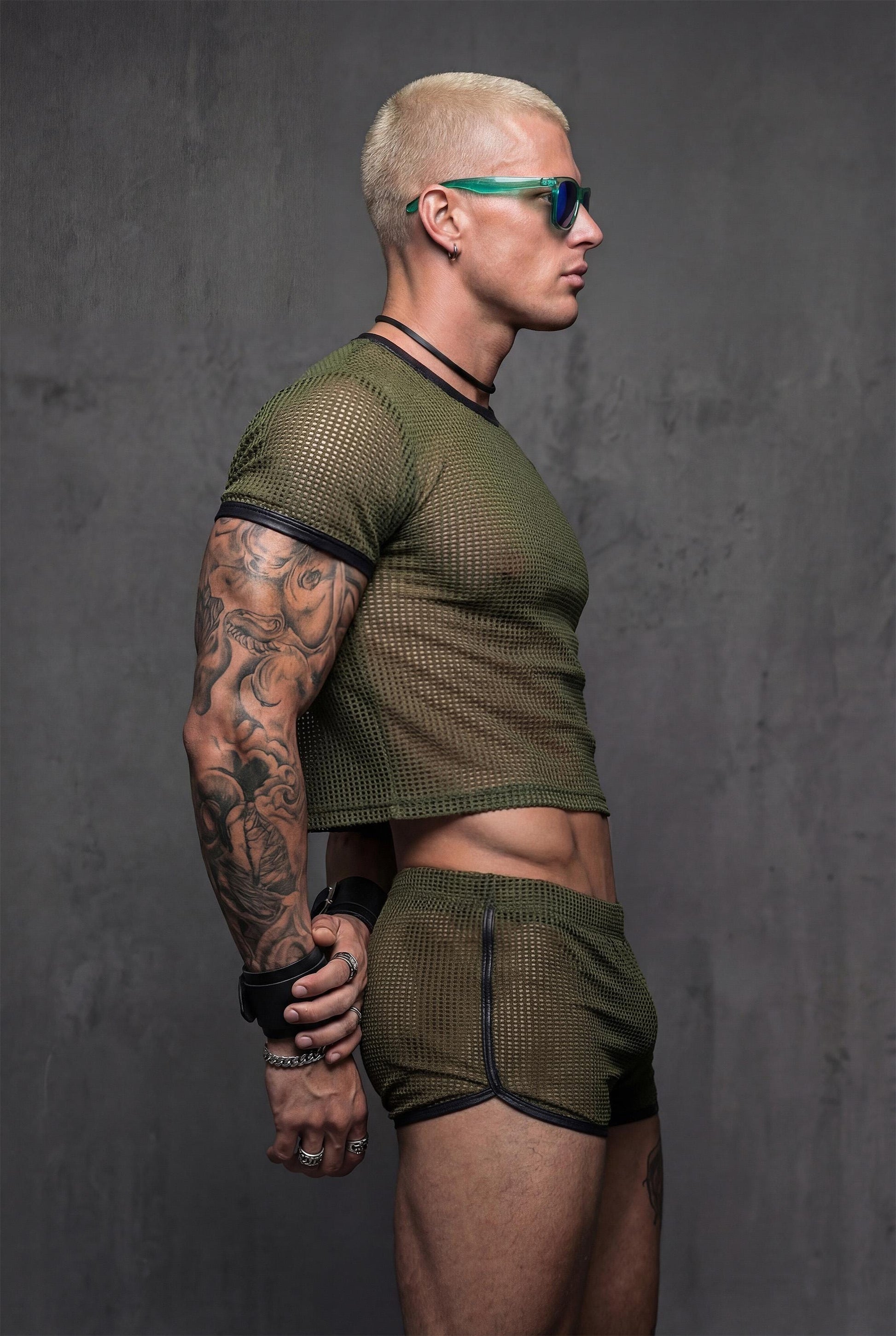 Side profile military green sheer mesh crop top with black vegan leather contrast trim paired with transparent shorts - PIEL Night Starter complete look for gay circuit parties underground raves - athletic body-hugging fit reveals masculine physique with confidence - queer menswear perfect for techno festivals pride parades fetish clubs beach parties - olive green alternative color stands out at rave events lgbtq celebrations