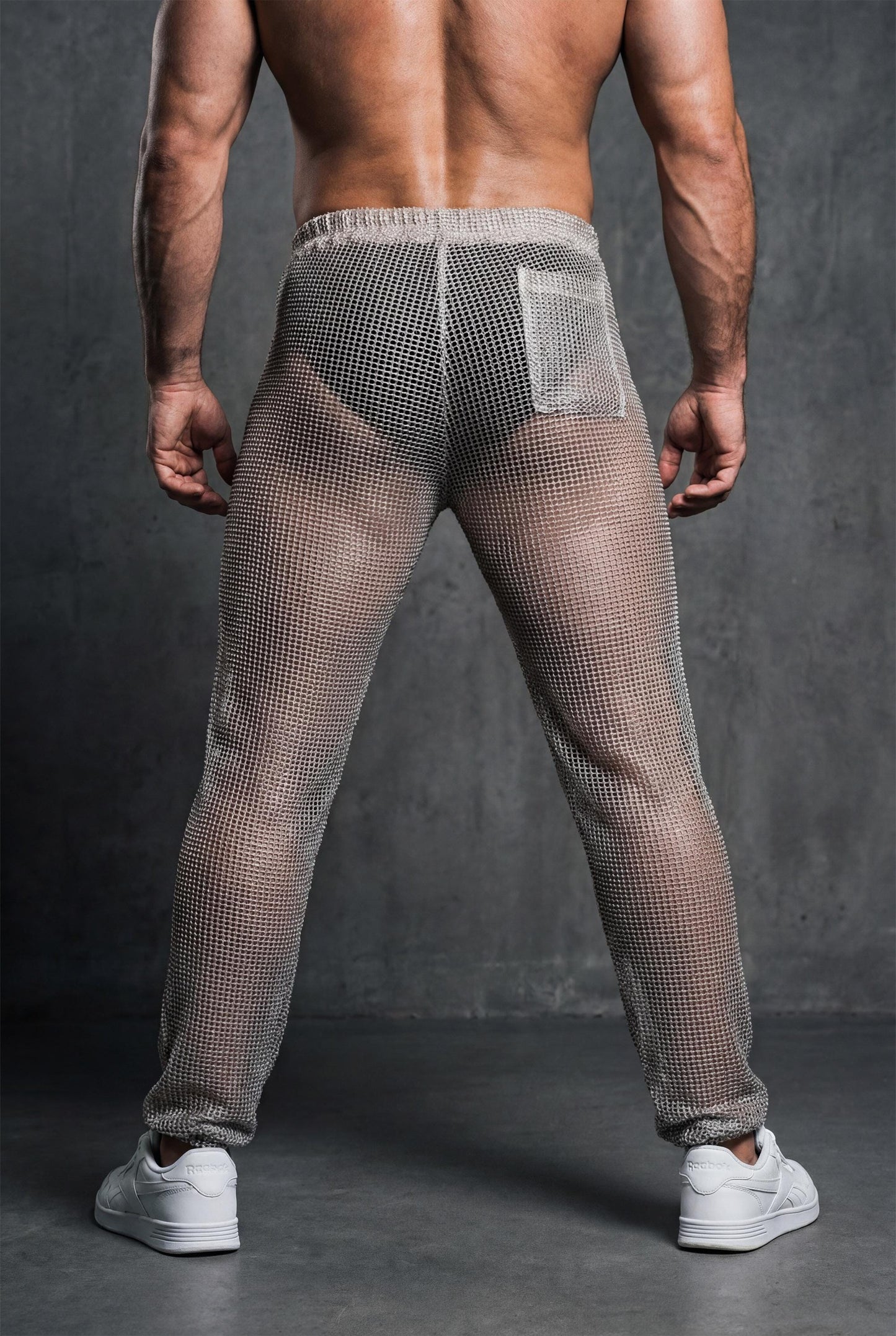 Beige see-through knit jogger pants men rear view showing loose tapered leg and breathable mesh texture for casual gay wear and festival style. Open-knit construction offers maximum ventilation for hot weather dancing and outdoor events. Elastic cuffs create clean finish at ankles. Ideal for circuit parties, rave festivals, pride parades, and beach clubs. Comfortable statement piece for men who want relaxed fit with bold transparent aesthetic and unapologetic style expression.