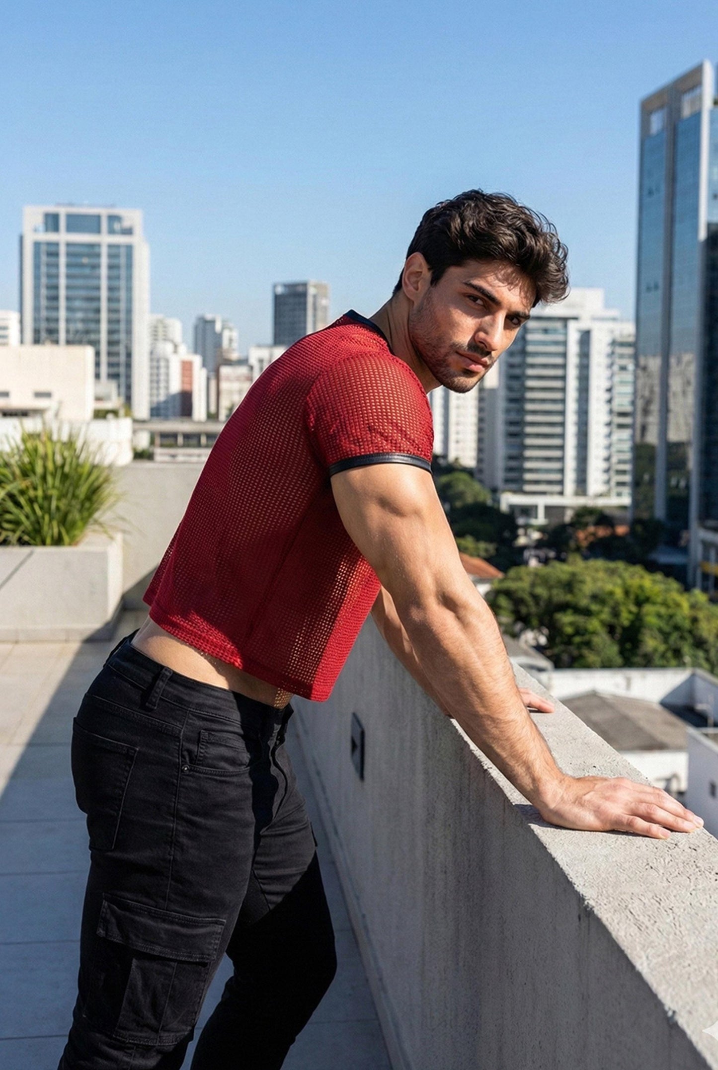 Muscular man wearing a red PIEL men’s mesh crop top on a rooftop with city skyline in the background. The short mesh top features a breathable open net fabric with a fitted cut that highlights the arms and shoulders. Men’s mesh crop top designed for summer wear, club outfits, rooftop parties, and bold masculine fashion. PIEL party top with confident, modern style.