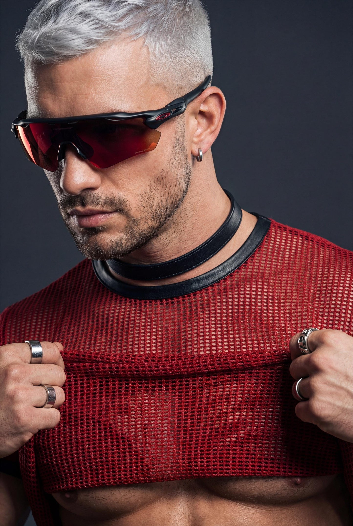 Muscular man wearing a red PIEL men’s mesh crop top in a studio close-up. The short mesh top features an open net texture with a fitted cut, revealing the chest while maintaining a structured neckline. PIEL men’s party top designed for club wear, nightlife outfits, and bold queer fashion aesthetics. Premium mesh top, confident masculine style.