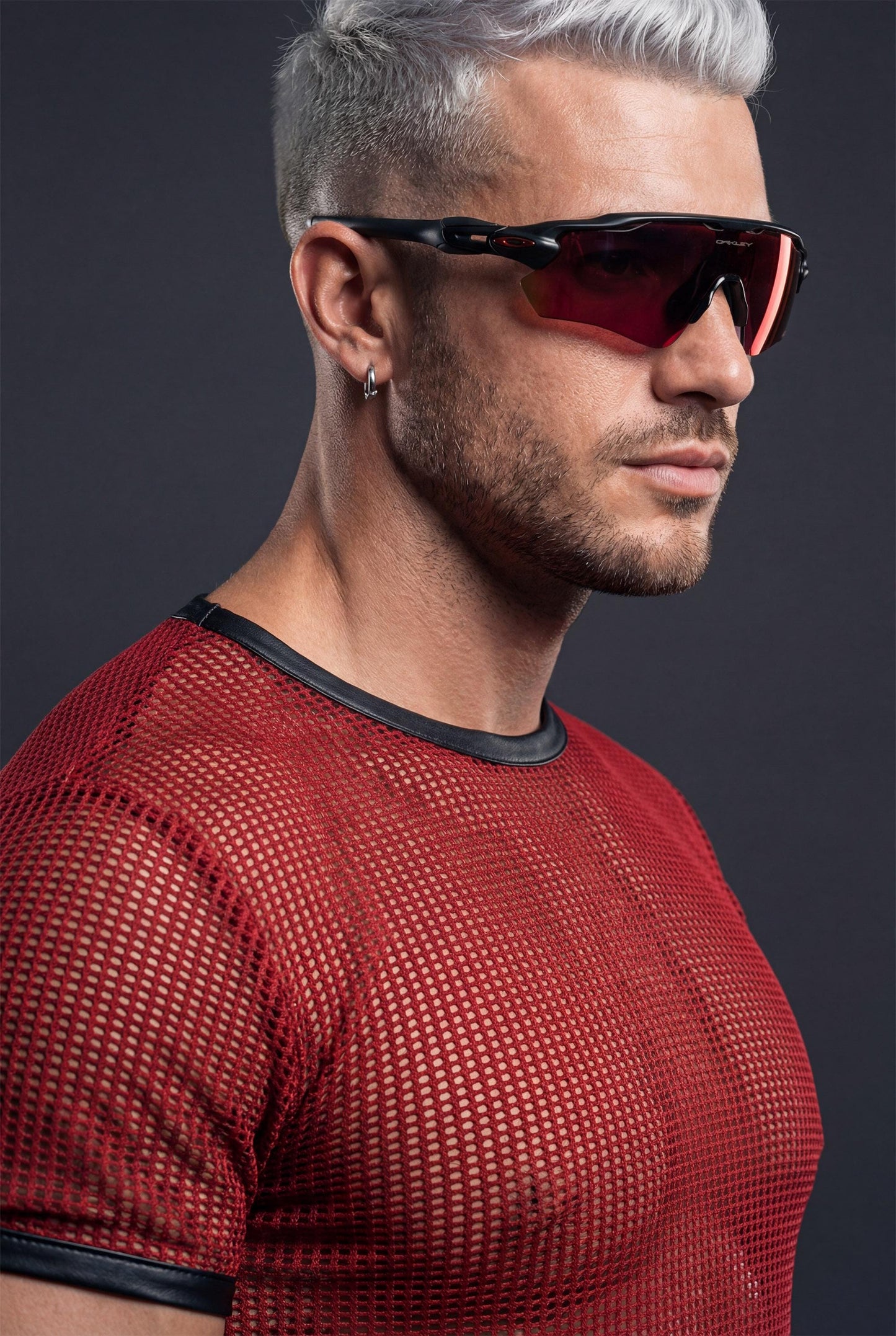 Muscular man wearing a red PIEL men’s mesh crop top in a studio close-up. The short mesh top features an open net texture with a fitted cut, revealing the chest while maintaining a structured neckline. PIEL men’s party top designed for club wear, nightlife outfits, and bold queer fashion aesthetics. Premium mesh top, confident masculine style.